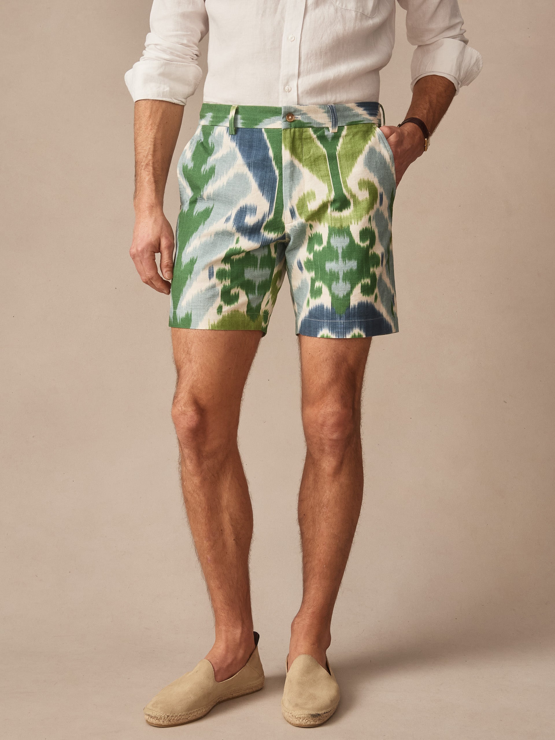A man models J.McLaughlin’s Chatham Shorts in Ikat, featuring a blue and green patterned design paired with a white shirt and beige slip-on shoes, showcasing effortlessly stylish, enduring designs and innovative fabrics in a legacy of timeless fashion. A destination for defining style. Life, Well-Lived.