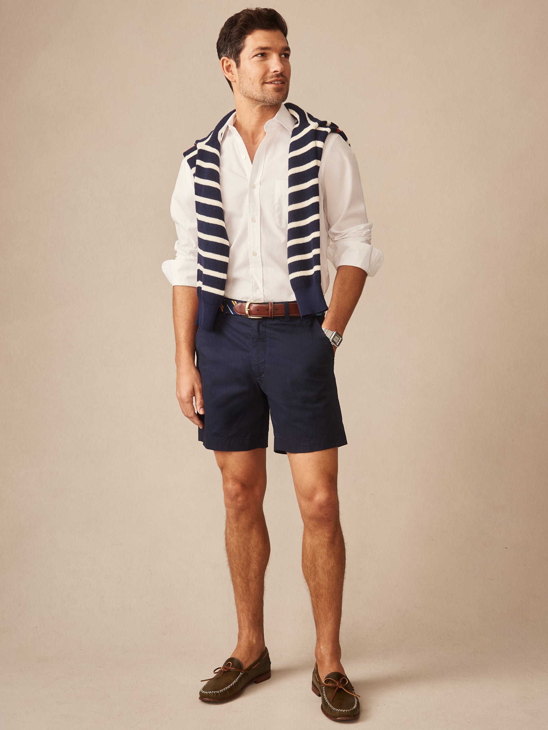 Man wearing navy Chatham Shorts by J.McLaughlin paired with a white button-down shirt, brown belt, striped sweater draped over shoulders, and casual loafers, showcasing effortlessly stylish, enduring designs and innovative fabrics from a legacy of timeless fashion. A destination for defining style. Life, Well-Lived.