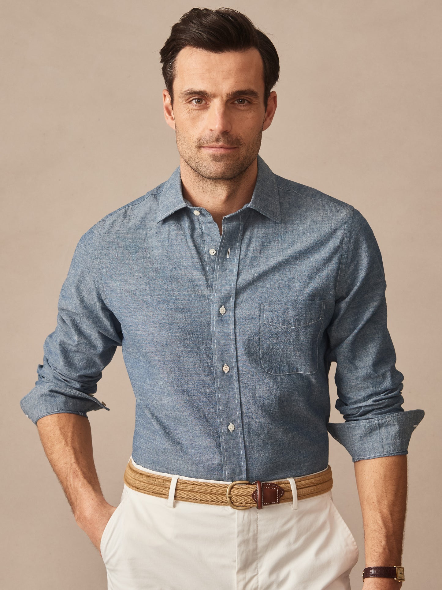 Man wearing a Gramercy Cotton Chambray Shirt by J.McLaughlin in a classic blue shade paired with cream pants and a tan belt, showcasing effortlessly stylish, enduring designs.