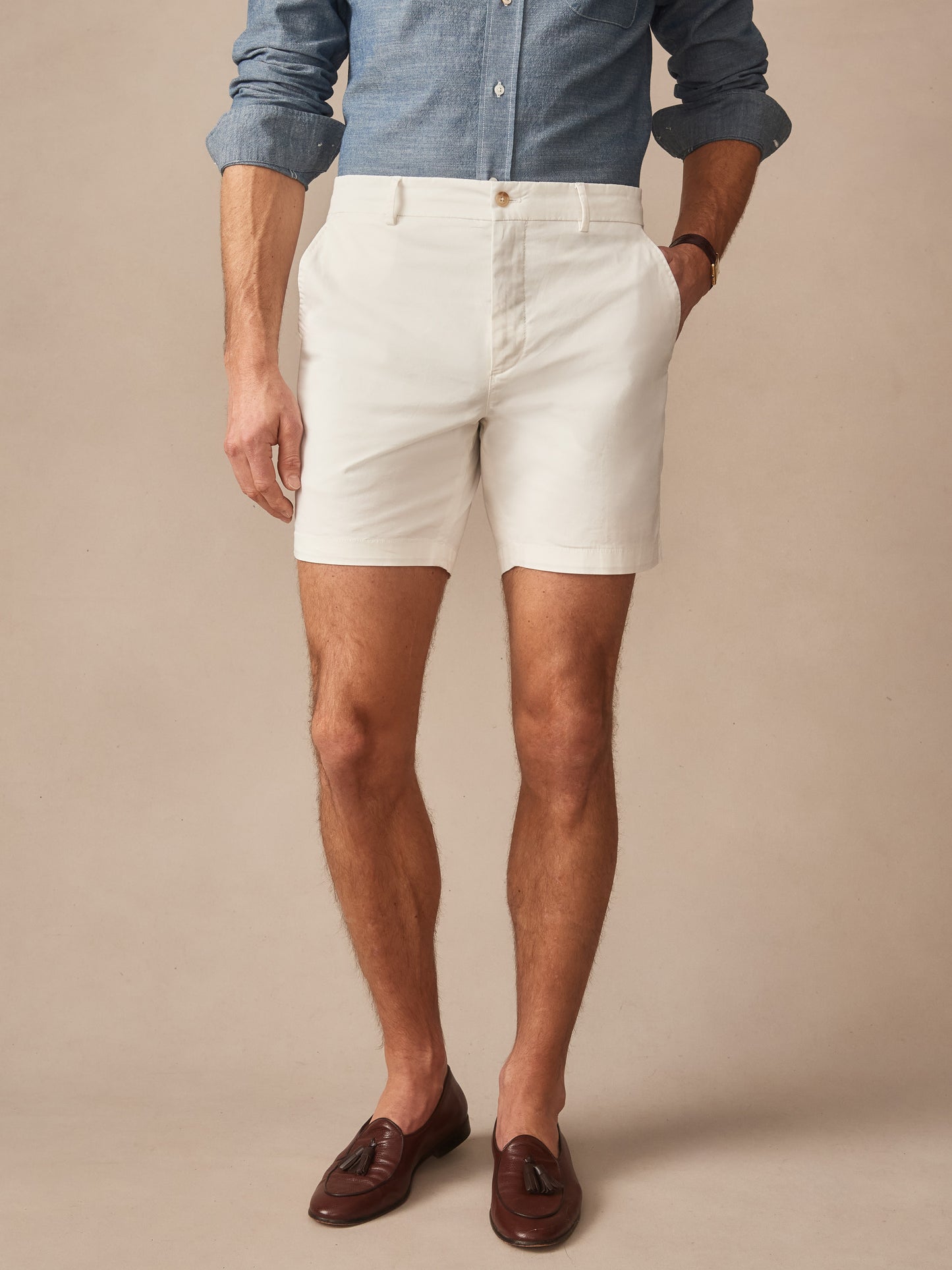 A man wearing J.McLaughlin Chatham Shorts in a classic stone color paired with a casual blue shirt and brown loafers, showcasing effortlessly stylish shorts with enduring designs and innovative fabrics.