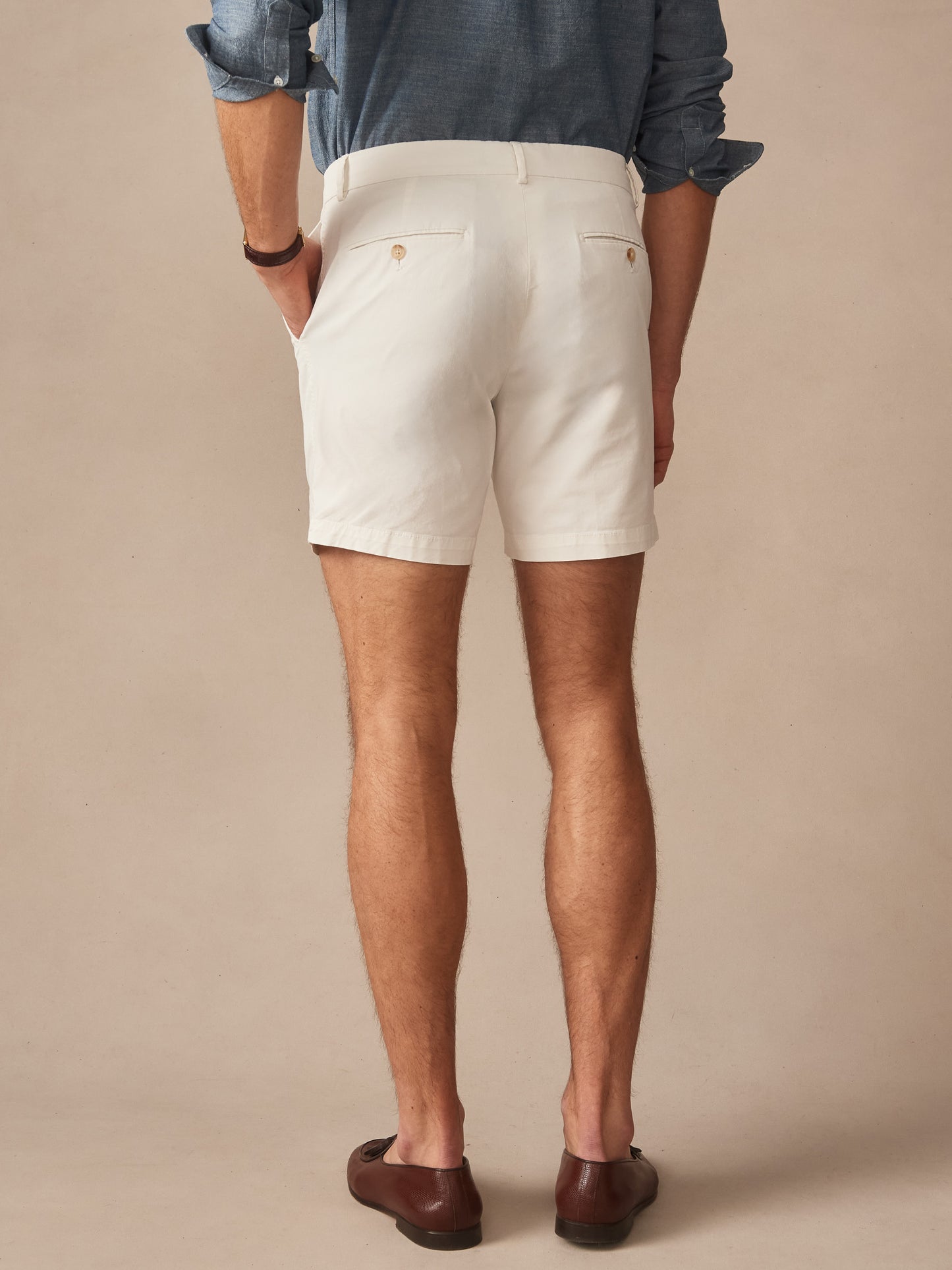 Rear view of a man wearing J.McLaughlin Chatham Shorts in stone color, paired with a rolled-up denim shirt and brown loafers, showcasing effortlessly stylish, enduring designs with a legacy of timeless fashion. A destination for defining style. Life, Well-Lived.