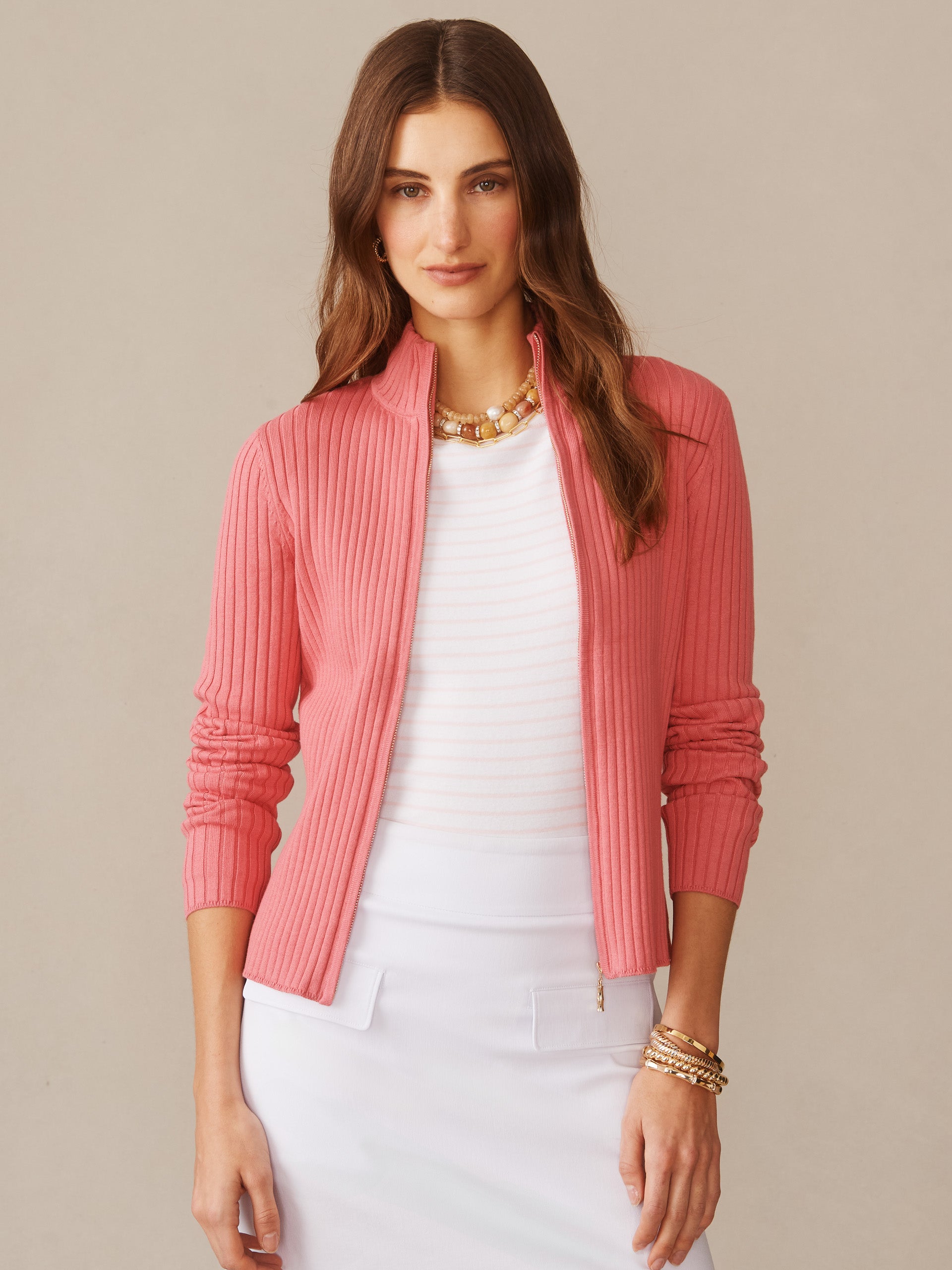 A woman wearing a pink Clemence Zip Cardigan by J.McLaughlin over a white top, styled with layered bracelets and a statement necklace, showcasing effortlessly stylish, enduring designs in innovative fabrics. A destination for defining style. Life, Well-Lived.