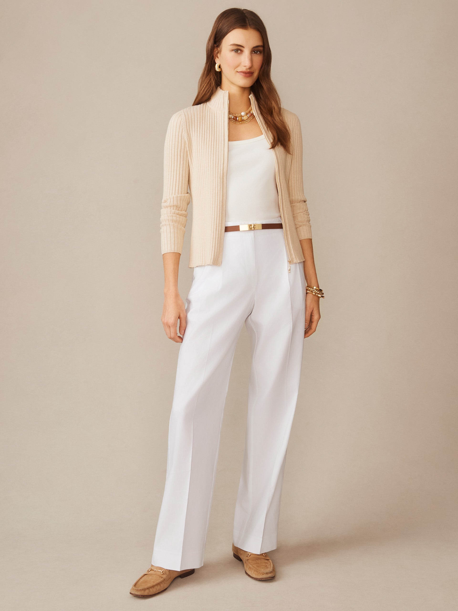 Woman wearing a beige Clemence Zip Cardigan by J.McLaughlin, styled with white pants and gold accessories, showcasing effortlessly stylish, enduring designs in a timeless look.
