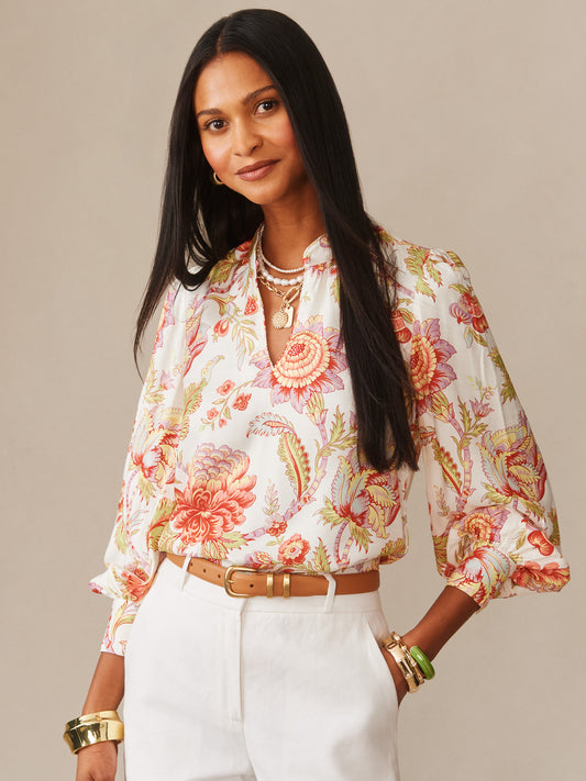 Woman wearing the Maeve Silk Blend Blouse in Cerulean Garden by J.McLaughlin, featuring a floral print with long sleeves, paired with white pants and gold and green accessories, embodying effortlessly stylish, enduring designs with innovative fabrics for a legacy of timeless fashion. A destination for defining style. Life, Well-Lived.