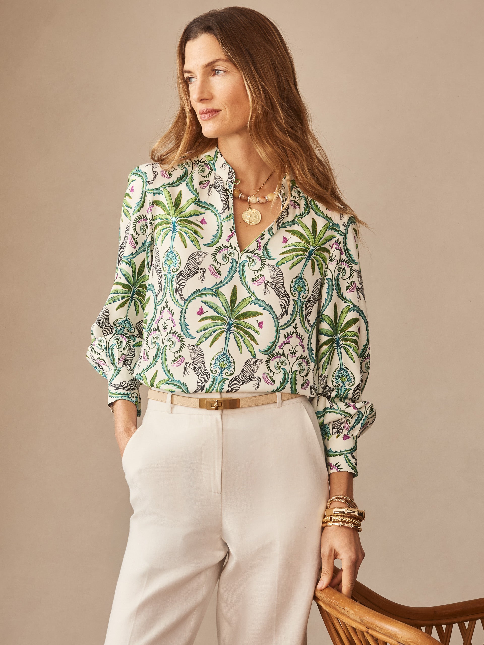 Woman wearing a Clover Silk Blend Blouse in Zebra Isle by J.McLaughlin featuring a vibrant palm tree and zebra print, paired with white trousers and gold accessories, embodying effortlessly stylish, enduring designs with innovative fabrics, a destination for defining style. Life, Well-Lived.