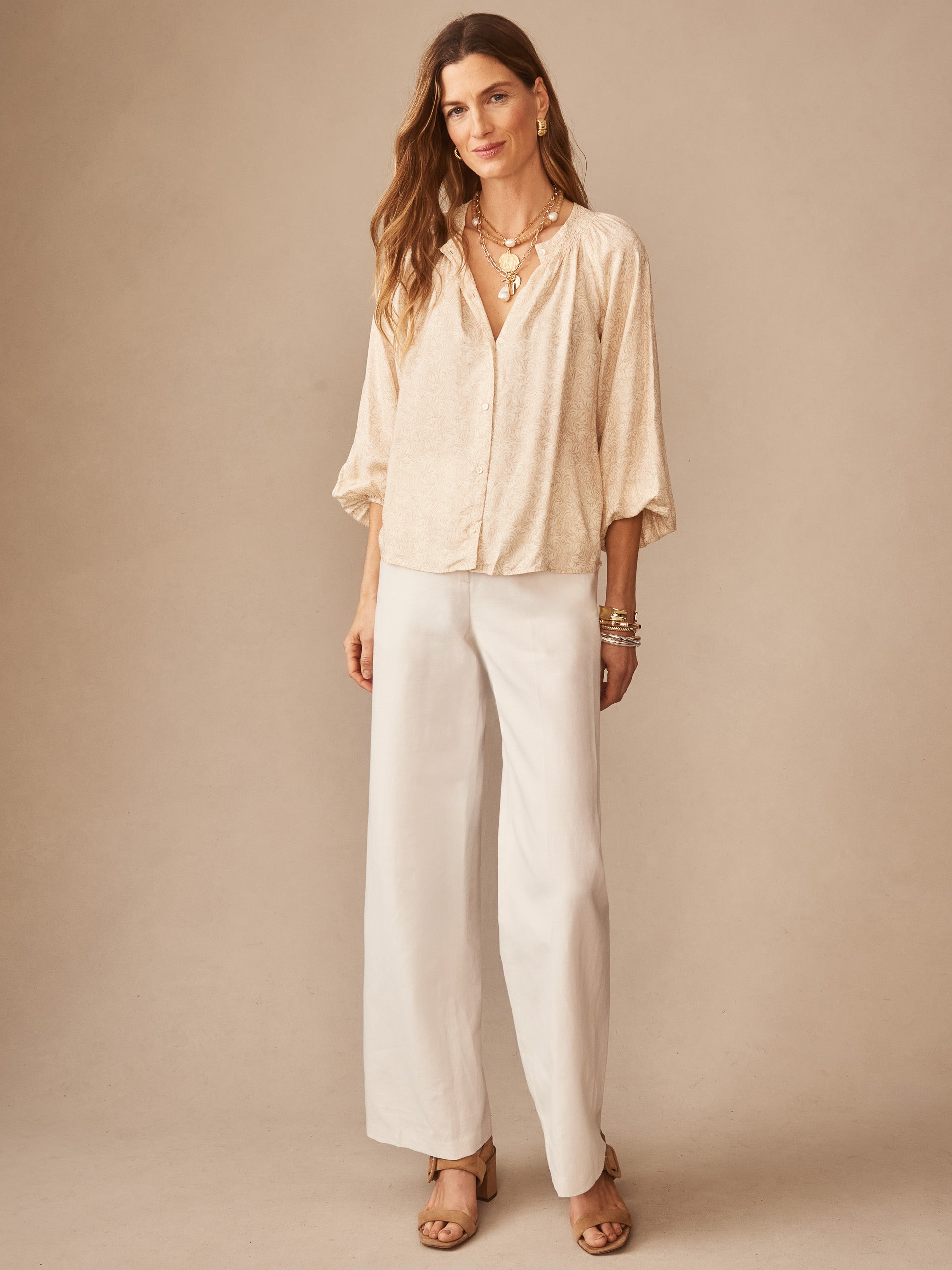 Woman wearing J.McLaughlin Constance Silk Blend Blouse in Mini Drawing Room, paired with wide-leg white pants and layered gold necklaces, showcasing effortlessly stylish, enduring designs.