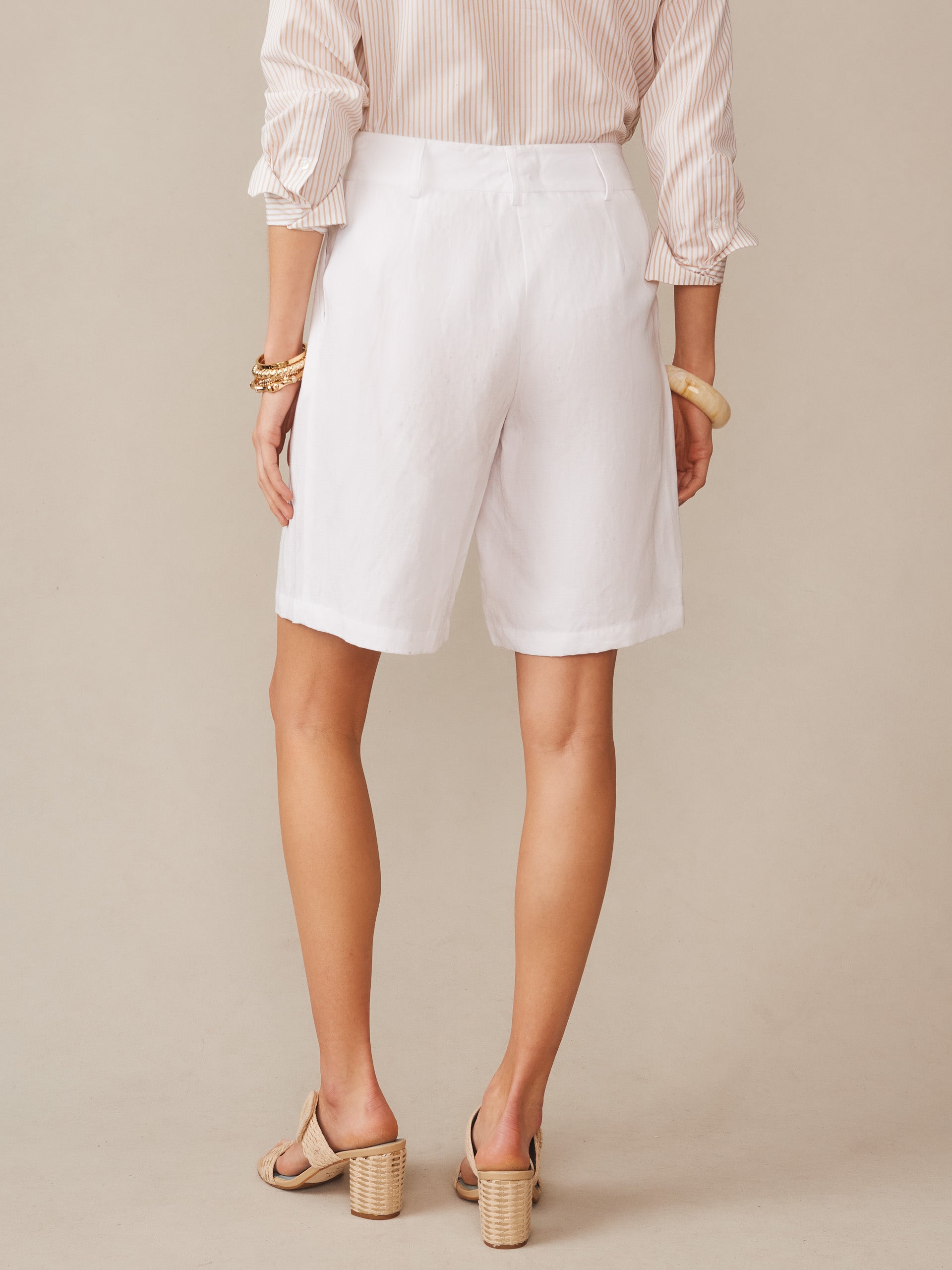 A woman wearing J.McLaughlin Cooper Linen Blend Shorts in white, styled with a striped blouse and heeled sandals, showcasing effortlessly stylish and enduring designs for a life well-lived.