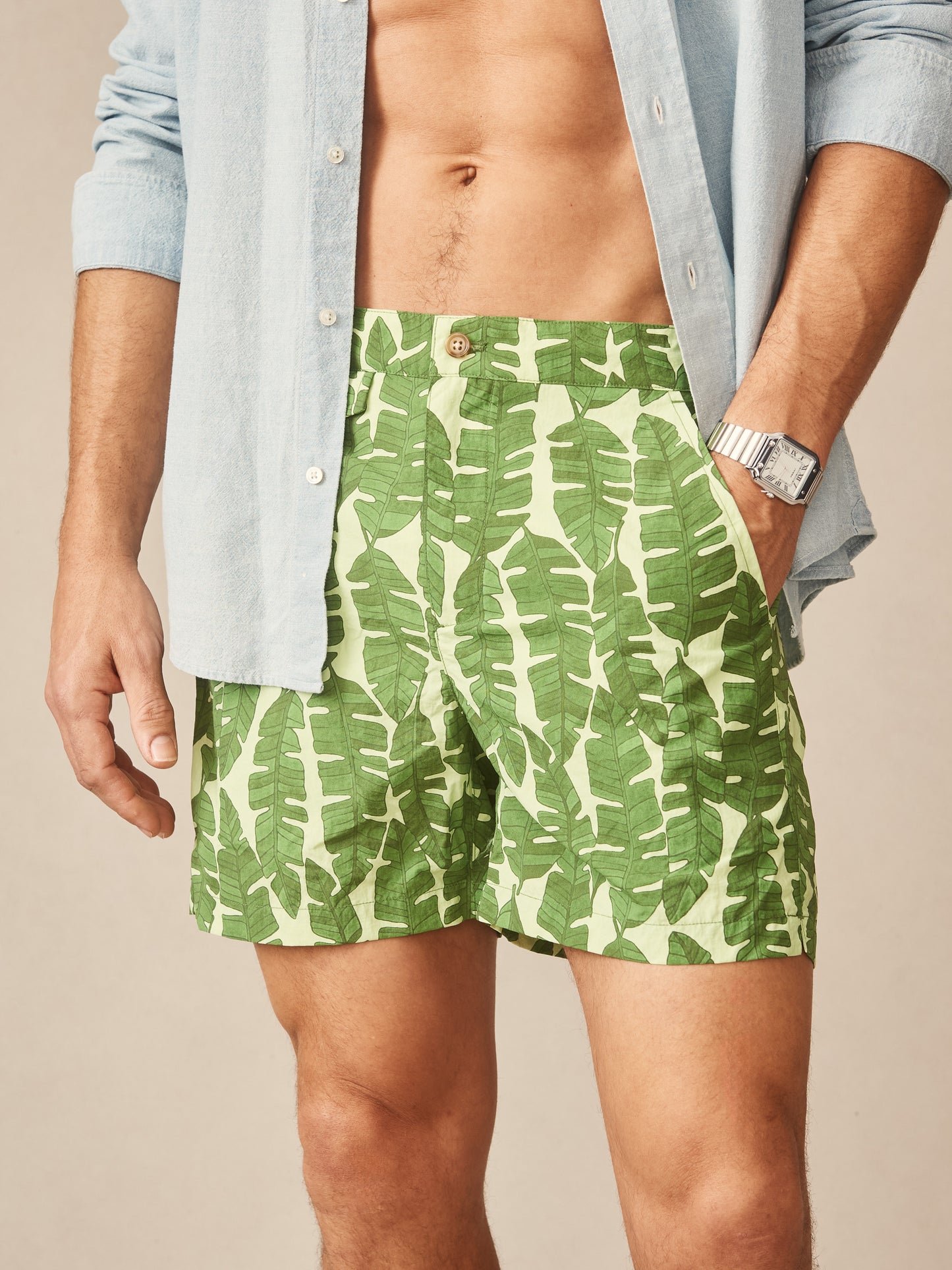 Close-up of a man wearing Deck Swim Trunks in Banana Leaves by J.McLaughlin with a green leafy print, paired with a light blue button-up shirt and a silver wristwatch.