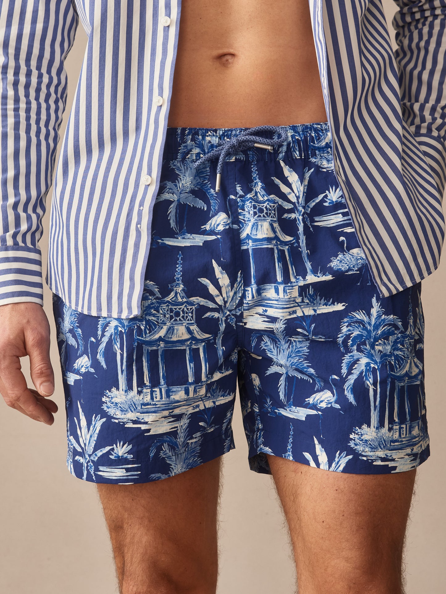 A close-up of a man wearing Marina Swim Trunks in Parisian Pergola by J.McLaughlin, featuring a blue tropical print with white and light blue pergola and palm tree motifs, paired with a blue and white striped shirt, showcasing effortlessly stylish swimwear with enduring designs and innovative fabrics, embodying a legacy of timeless fashion from a destination for defining style. Life, Well-Lived.