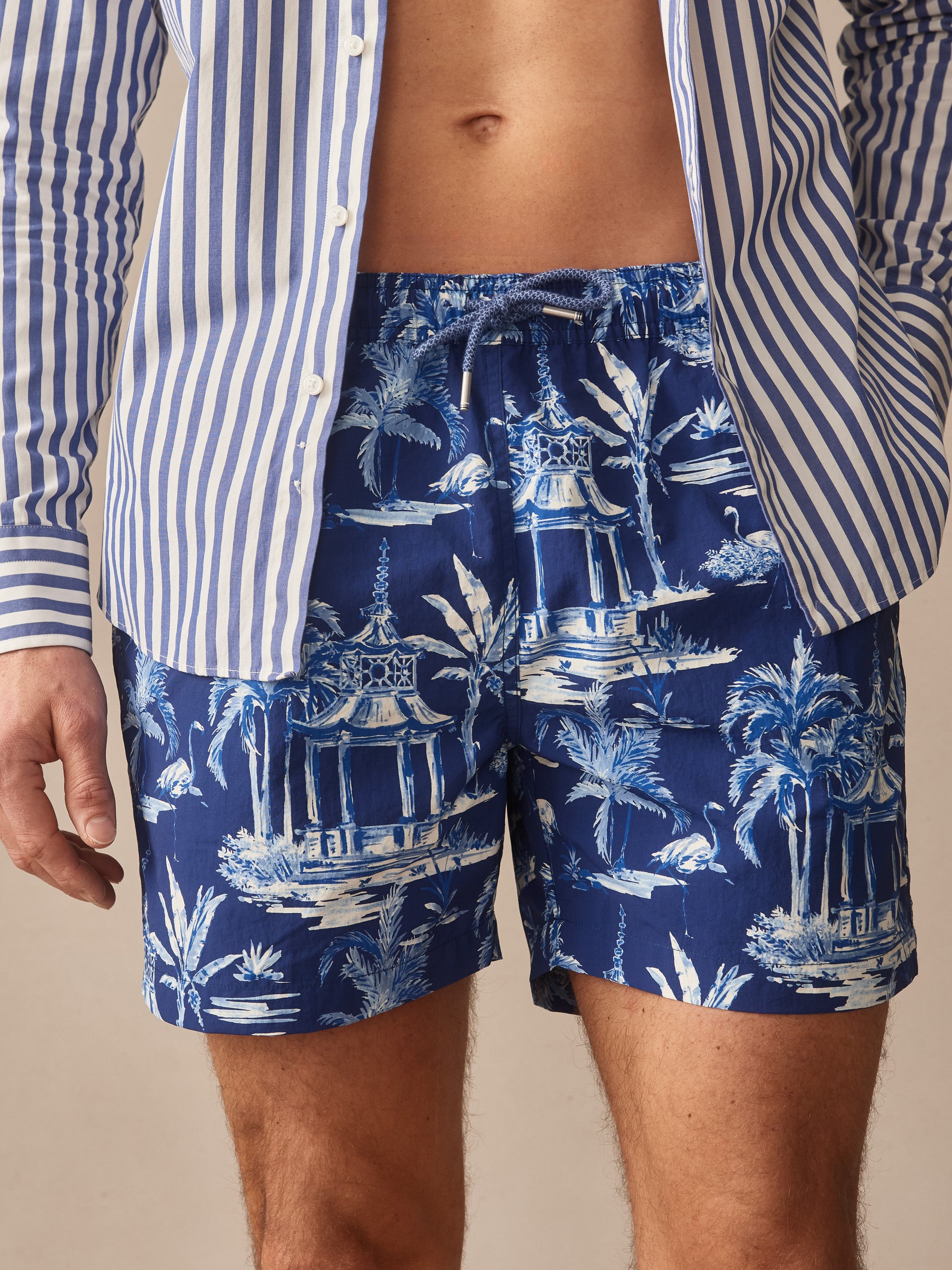 A close-up of a man wearing Marina Swim Trunks in Parisian Pergola by J.McLaughlin, featuring a blue tropical print with white and light blue pergola and palm tree motifs, paired with a blue and white striped shirt, showcasing effortlessly stylish swimwear with enduring designs and innovative fabrics, embodying a legacy of timeless fashion from a destination for defining style. Life, Well-Lived.