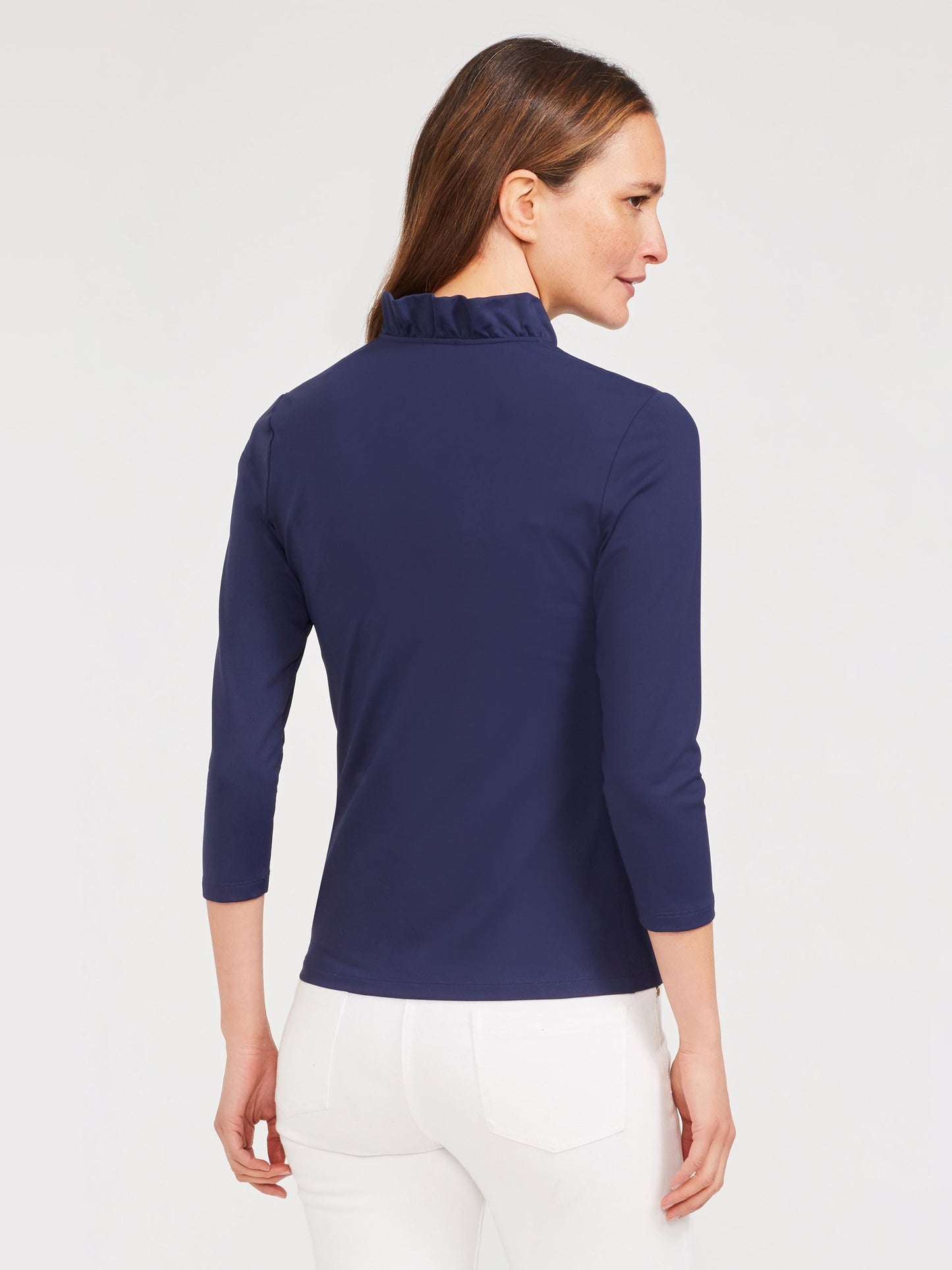Woman wearing a navy Durham Ruffle Top by J.McLaughlin, shown from the back with three-quarter sleeves and a subtle ruffle detail on the collar, paired with white pants.