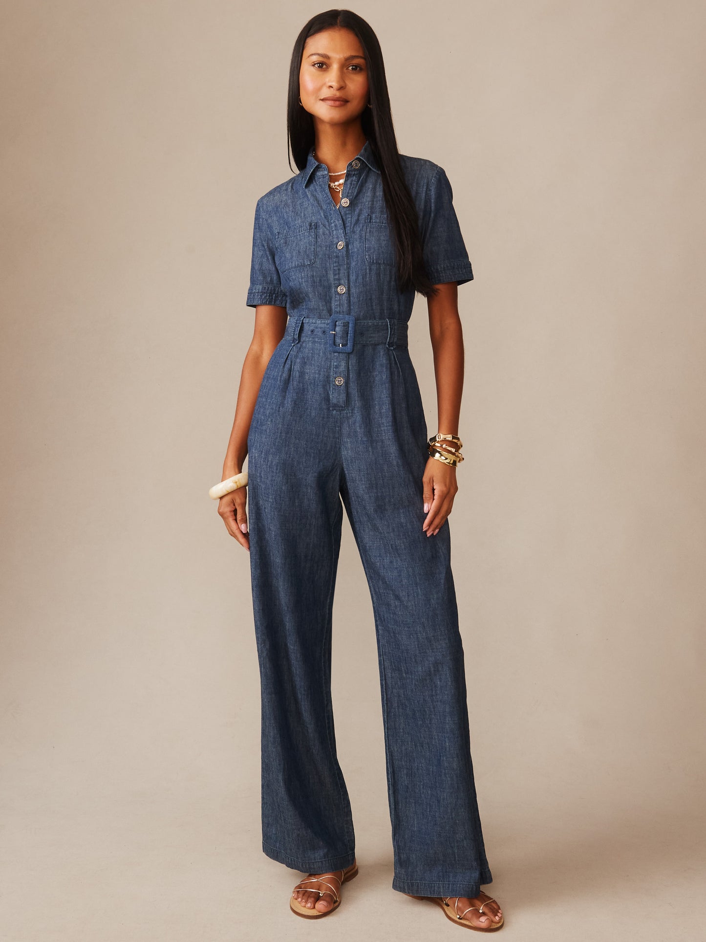 Woman wearing a denim Elena Jumpsuit by J.McLaughlin, featuring short sleeves, button front, belted waist, styled with bracelets and sandals, showcasing effortlessly stylish, enduring designs.