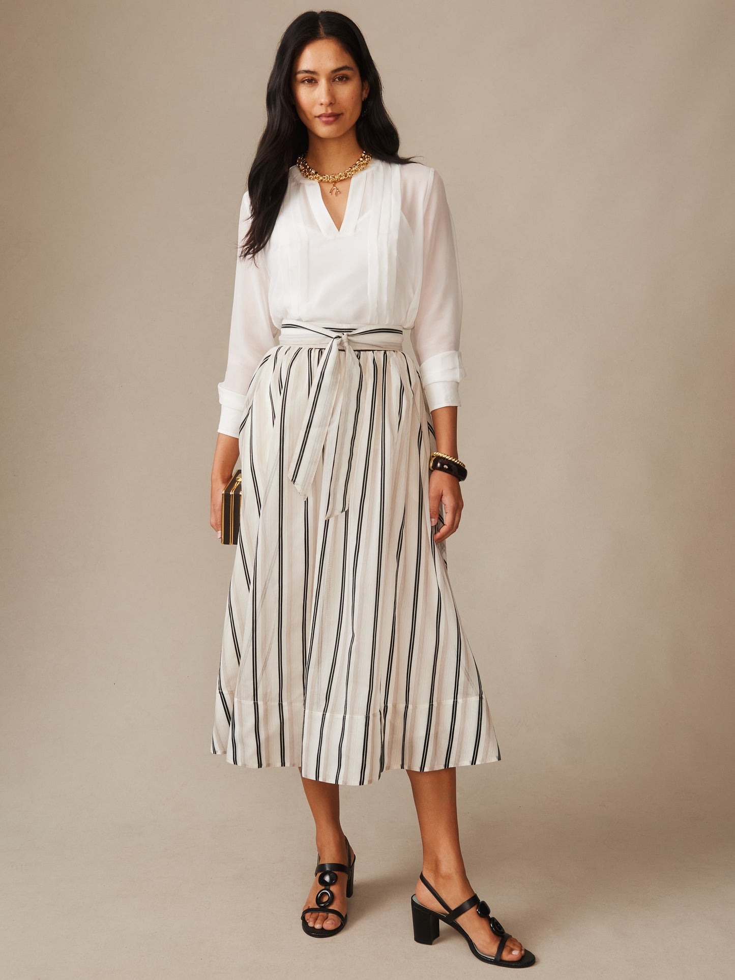 Woman wearing a white blouse and the Elenia Skirt in Stripe by J.McLaughlin, featuring vertical black stripes on a cream fabric, styled with black heeled sandals and gold accessories, showcasing effortlessly stylish, enduring designs in a timeless fashion look.