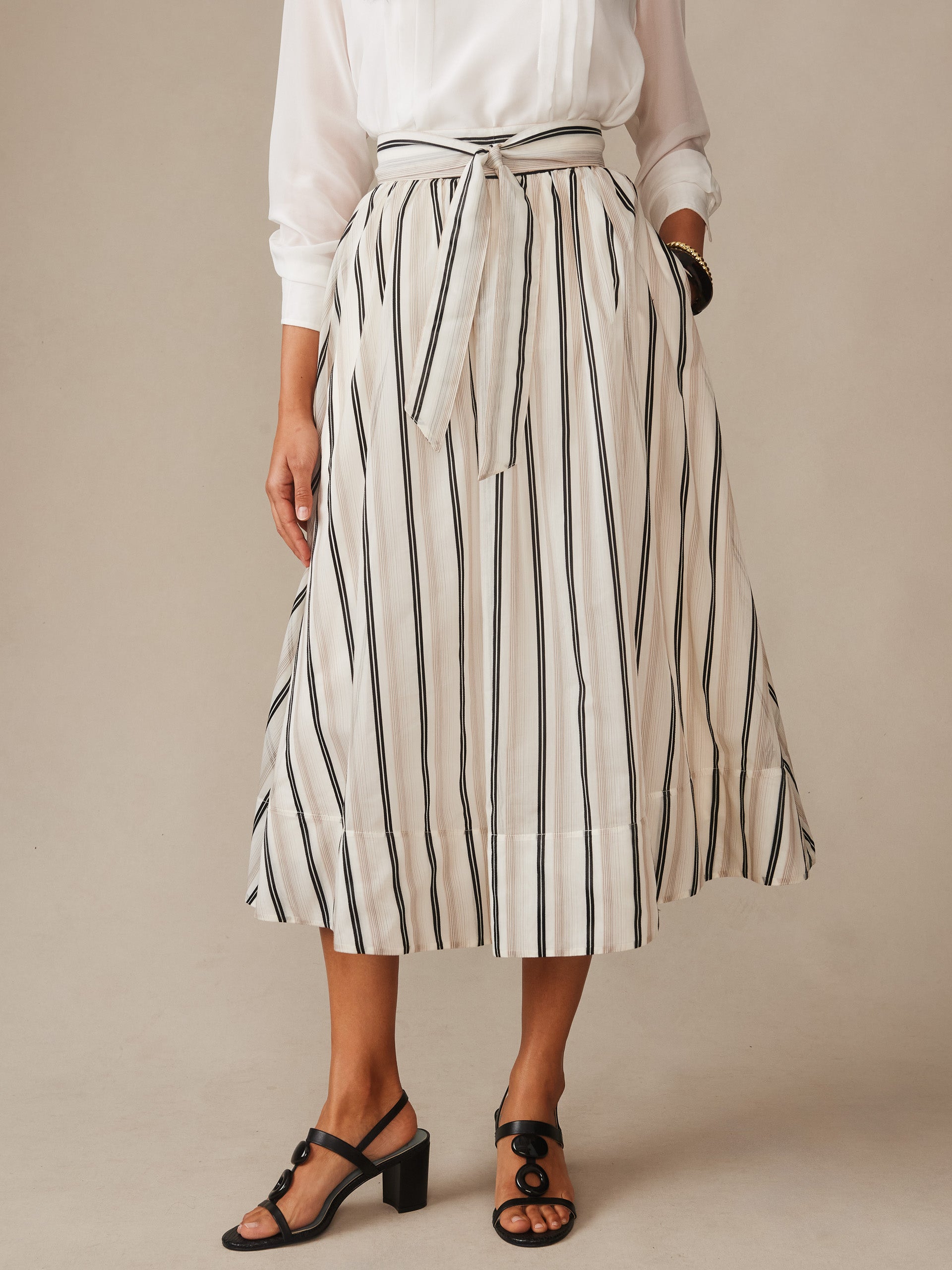 A classic J.McLaughlin Elenia Skirt in Stripe featuring cream fabric with vertical black stripes, paired with a white blouse and black heeled sandals, embodying effortlessly stylish, enduring designs and innovative fabrics with a legacy of timeless fashion. A destination for defining style. Life, Well-Lived.