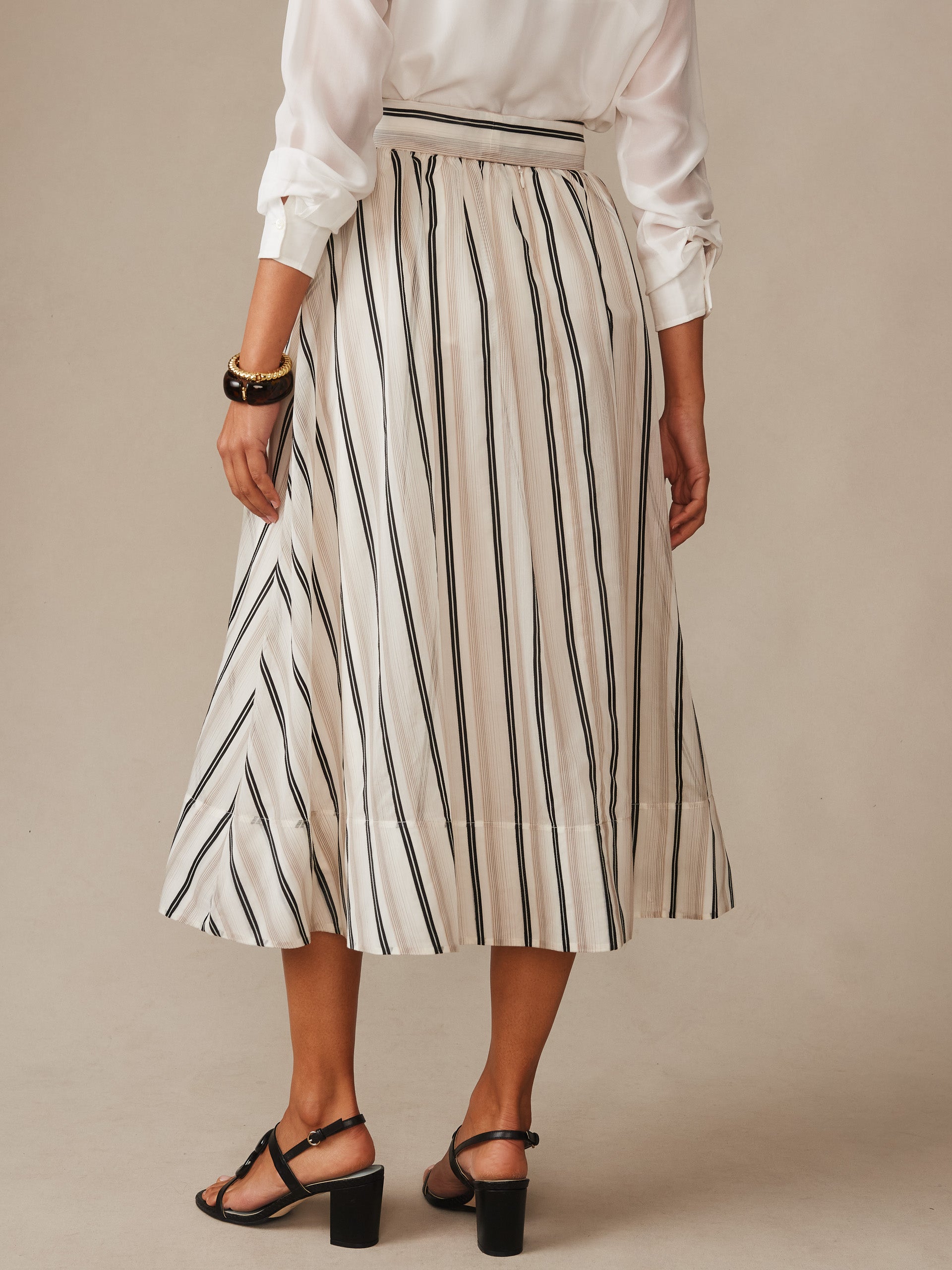 A woman modeling the Elenia Skirt in Stripe by J.McLaughlin, featuring a cream fabric with vertical black stripes, paired with a white blouse and black heels, showcasing effortlessly stylish, enduring designs with a legacy of timeless fashion. A destination for defining style. Life, Well-Lived.