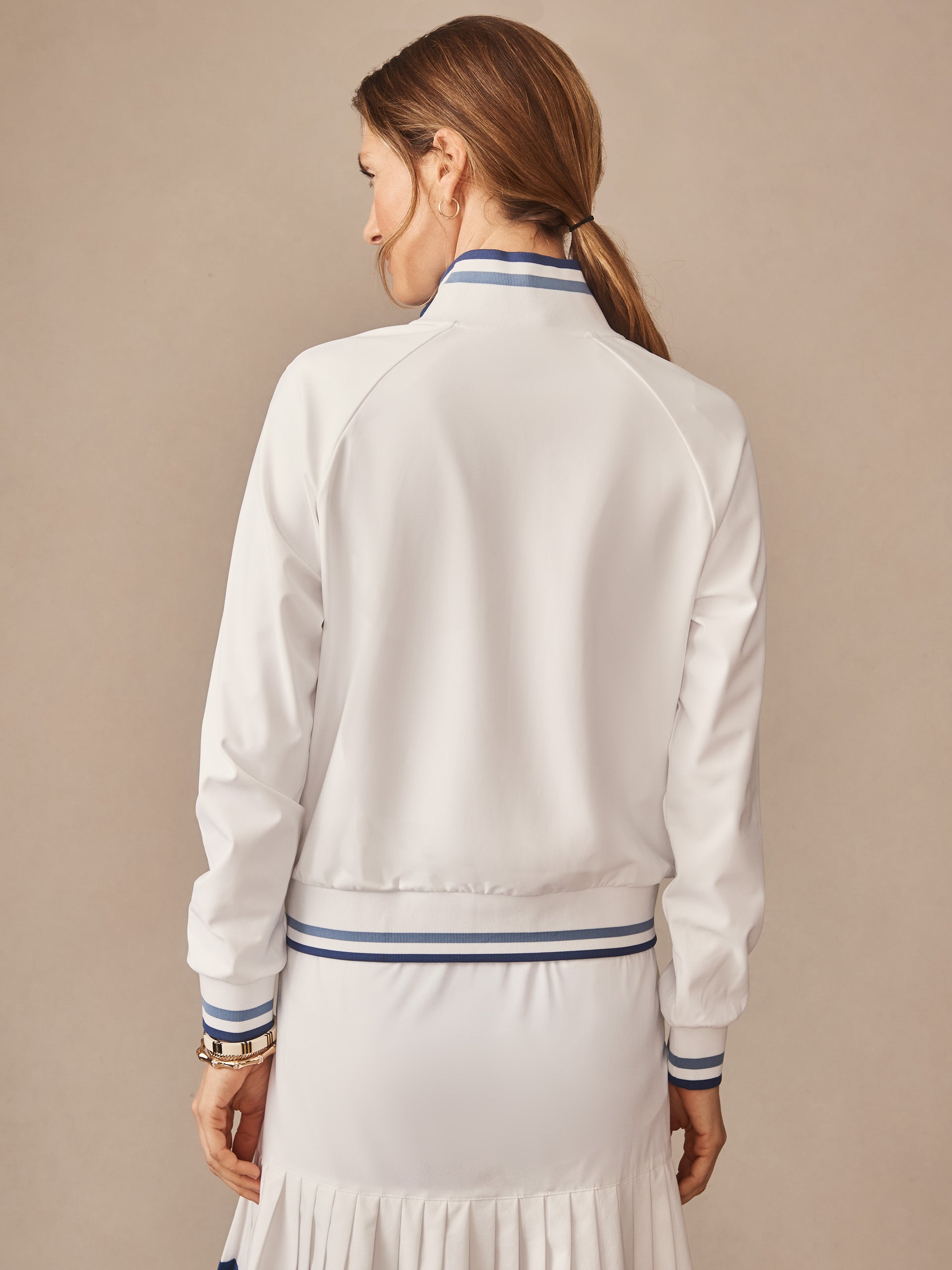 Woman wearing the back view of the white Elettra Jacket by J.McLaughlin featuring blue striped ribbed cuffs and waistband, showcasing effortlessly stylish, enduring designs with innovative fabrics.