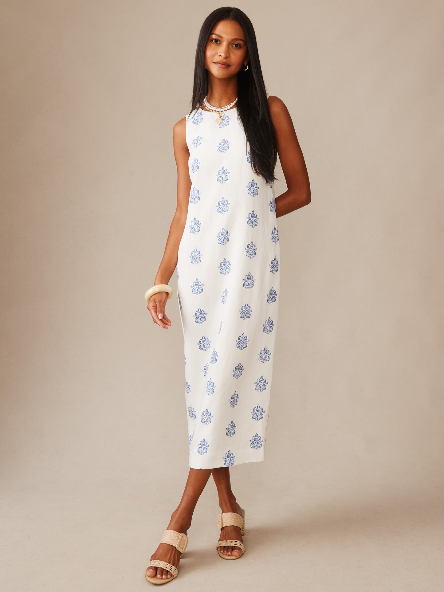 A woman modeling the Ella Linen Blend Dress in Apertivo by J.McLaughlin, featuring a white sleeveless design with blue patterns, styled with neutral sandals and elegant jewelry, showcasing effortlessly stylish and enduring designs crafted from innovative fabrics, embodying a legacy of timeless fashion. A destination for defining style. Life, Well-Lived.