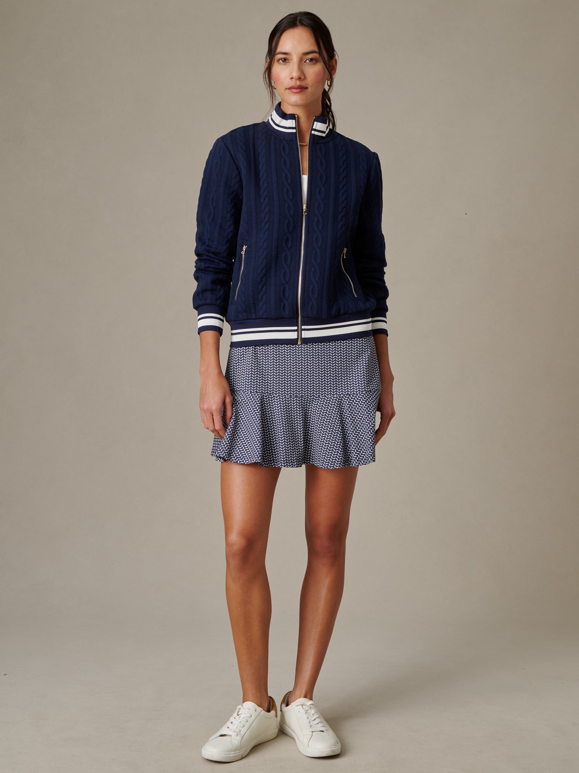 Woman wearing a navy Willa Jacket in Cable Jacquard by J.McLaughlin, styled with a patterned skirt and white sneakers, showcasing effortlessly stylish and enduring designs.