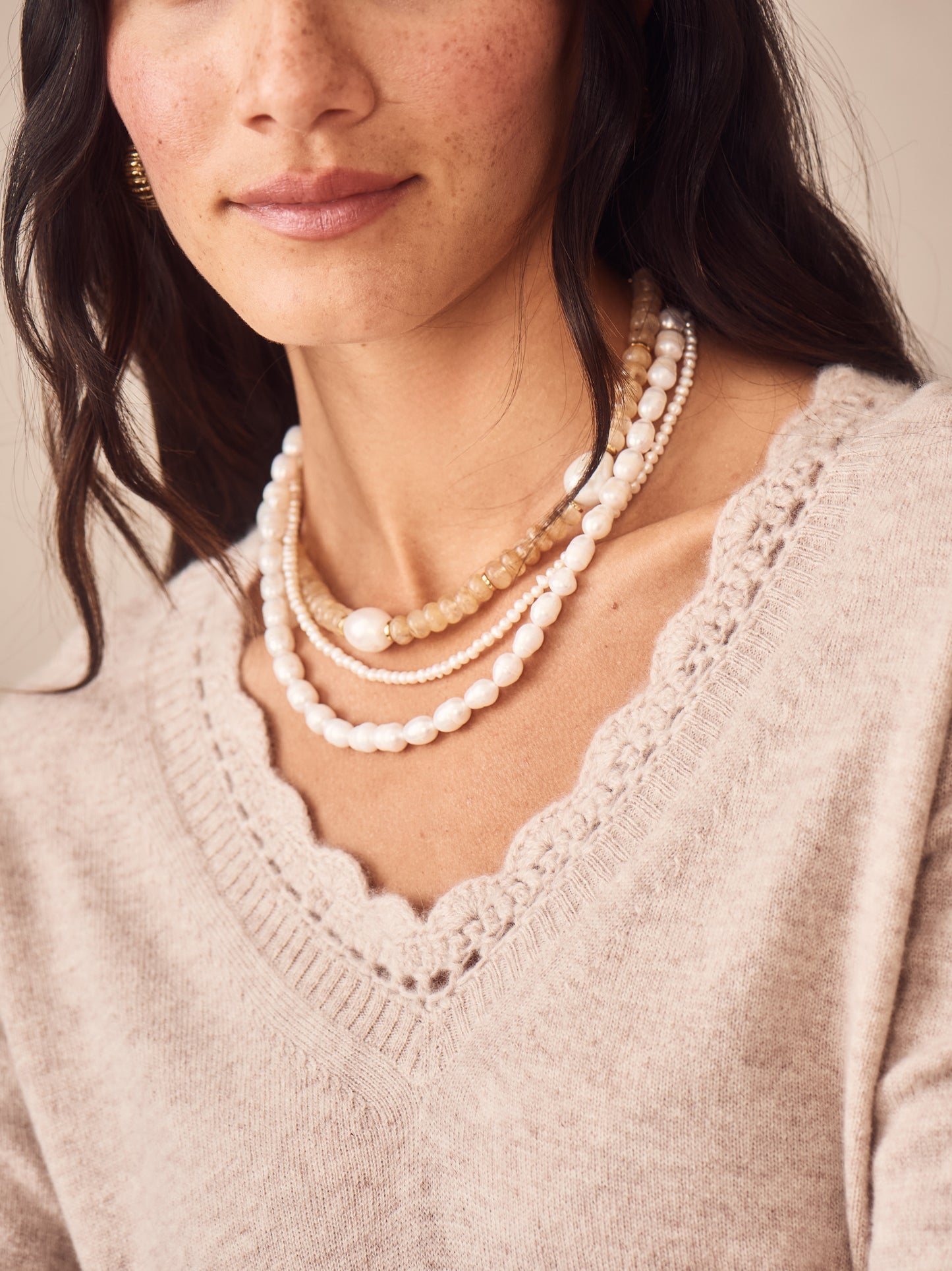 Close-up of a woman wearing an oatmeal Evelyn Cashmere Sweater by J.McLaughlin, showcasing its delicate scalloped V-neck design paired with layered neutral-tone beaded necklaces, embodying effortlessly stylish, enduring designs and a legacy of timeless fashion at a destination for defining style. Life, Well-Lived.