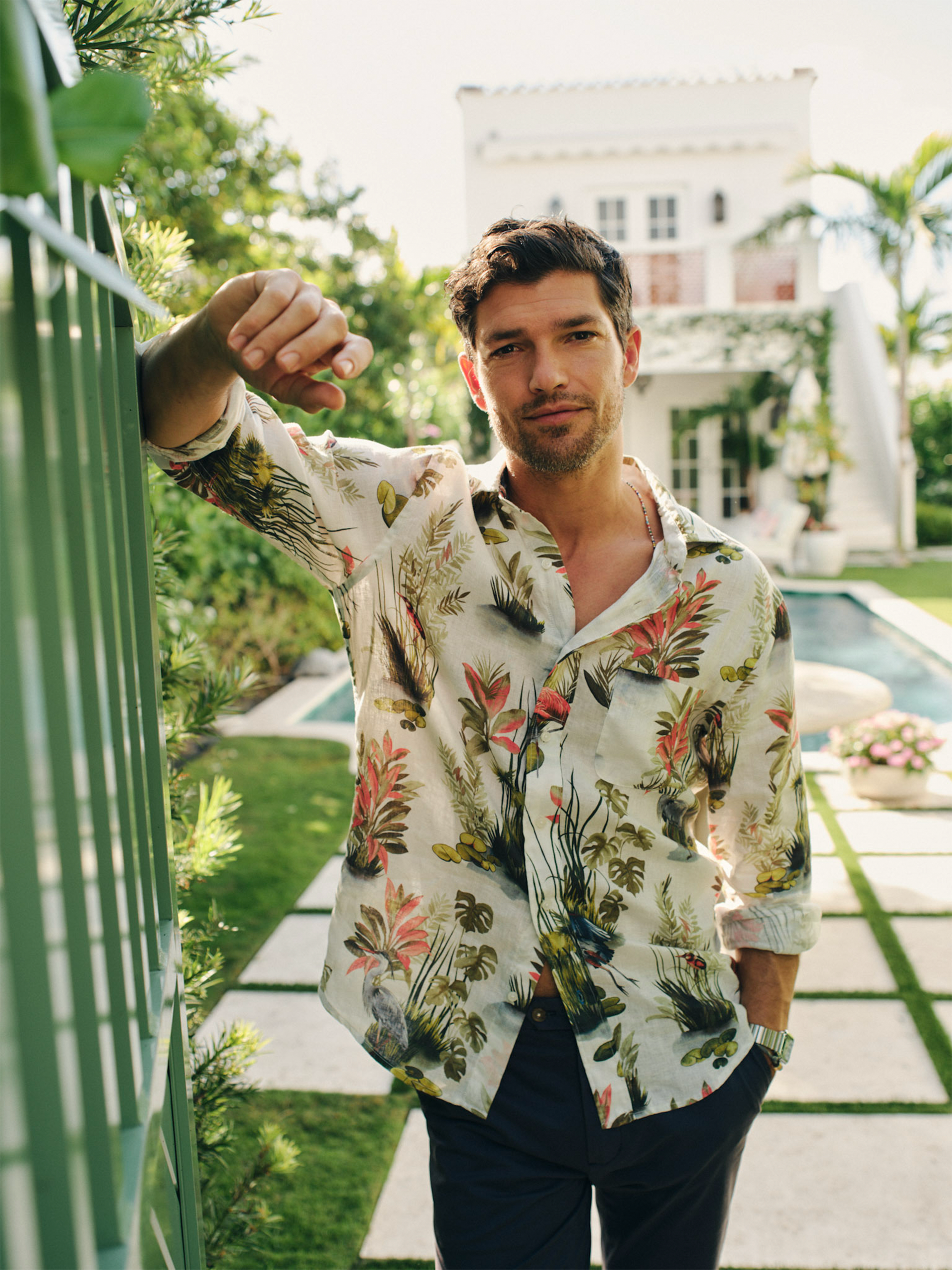 A man wearing the Gramercy Linen Shirt in Tidal Palms by J.McLaughlin, standing casually by a garden gate with greenery and a pool in the background, showcasing effortlessly stylish, enduring designs in innovative fabrics for a lifestyle well-lived.