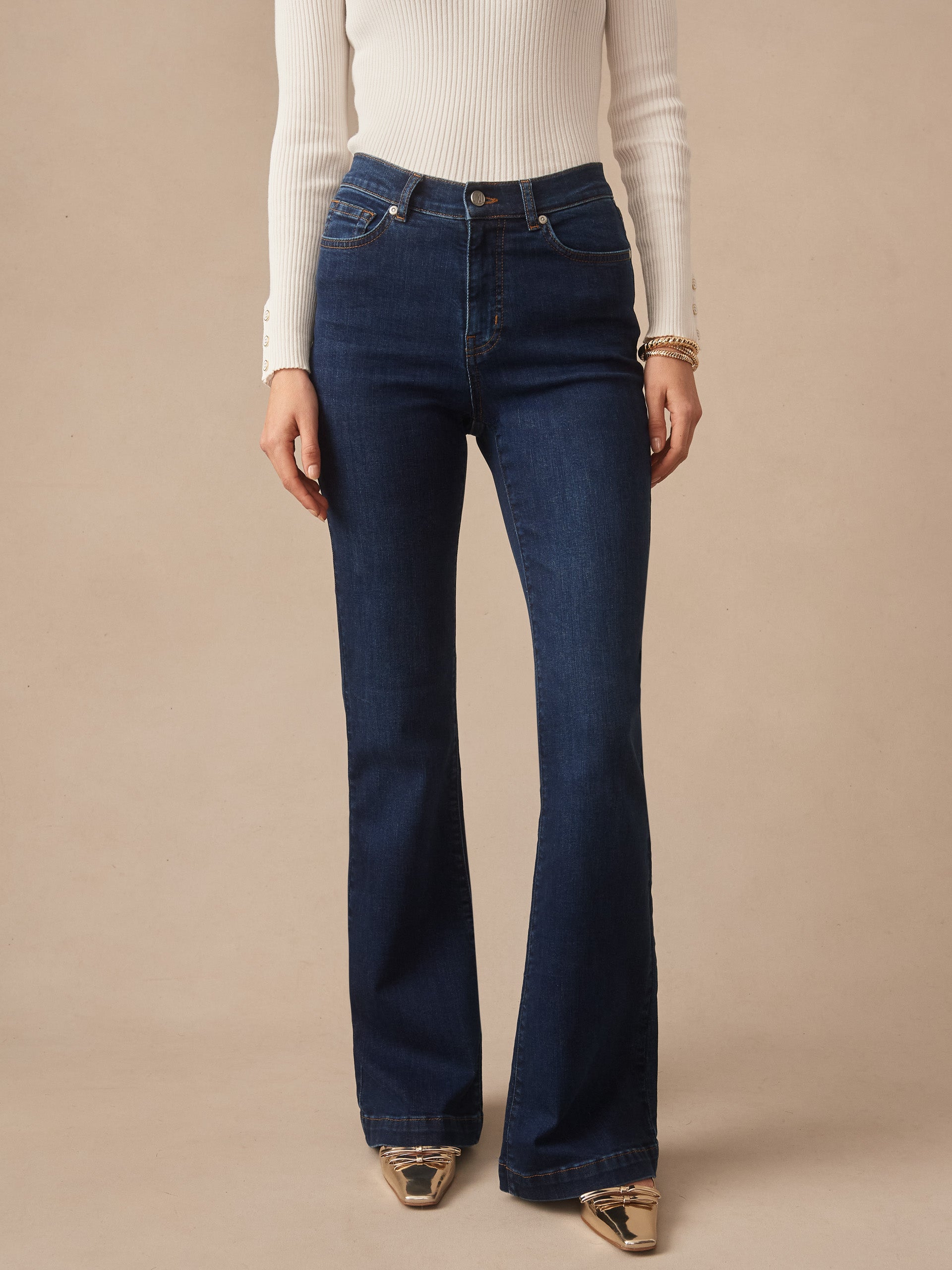 Woman wearing J.McLaughlin Domino Jeans in dark denim paired with a cream ribbed long-sleeve top and gold shoes, showcasing effortlessly stylish and enduring designs.