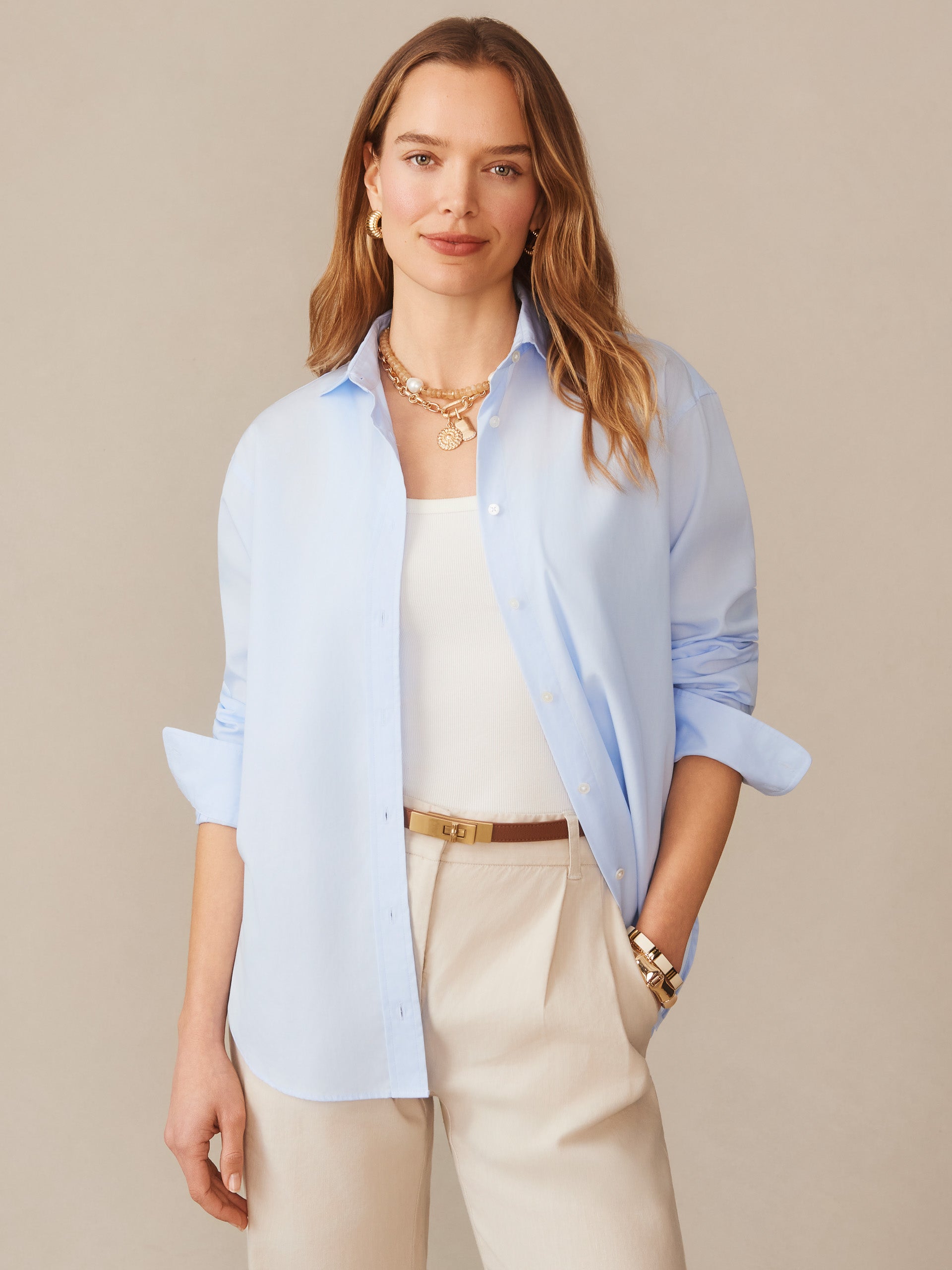 A woman wearing the Finn Shirt by J.McLaughlin in light blue, styled with neutral pants and layered gold jewelry, showcasing effortlessly stylish, enduring designs with innovative fabrics in a legacy of timeless fashion. A destination for defining style. Life, Well-Lived.