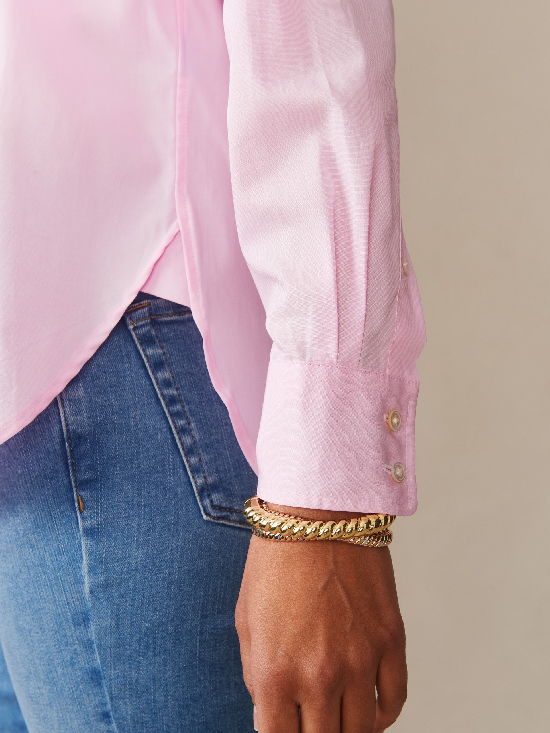 Close-up of a person wearing a soft pink Finn Shirt by J.McLaughlin paired with blue denim, accessorized with elegant gold bracelets, reflecting effortlessly stylish and enduring designs.