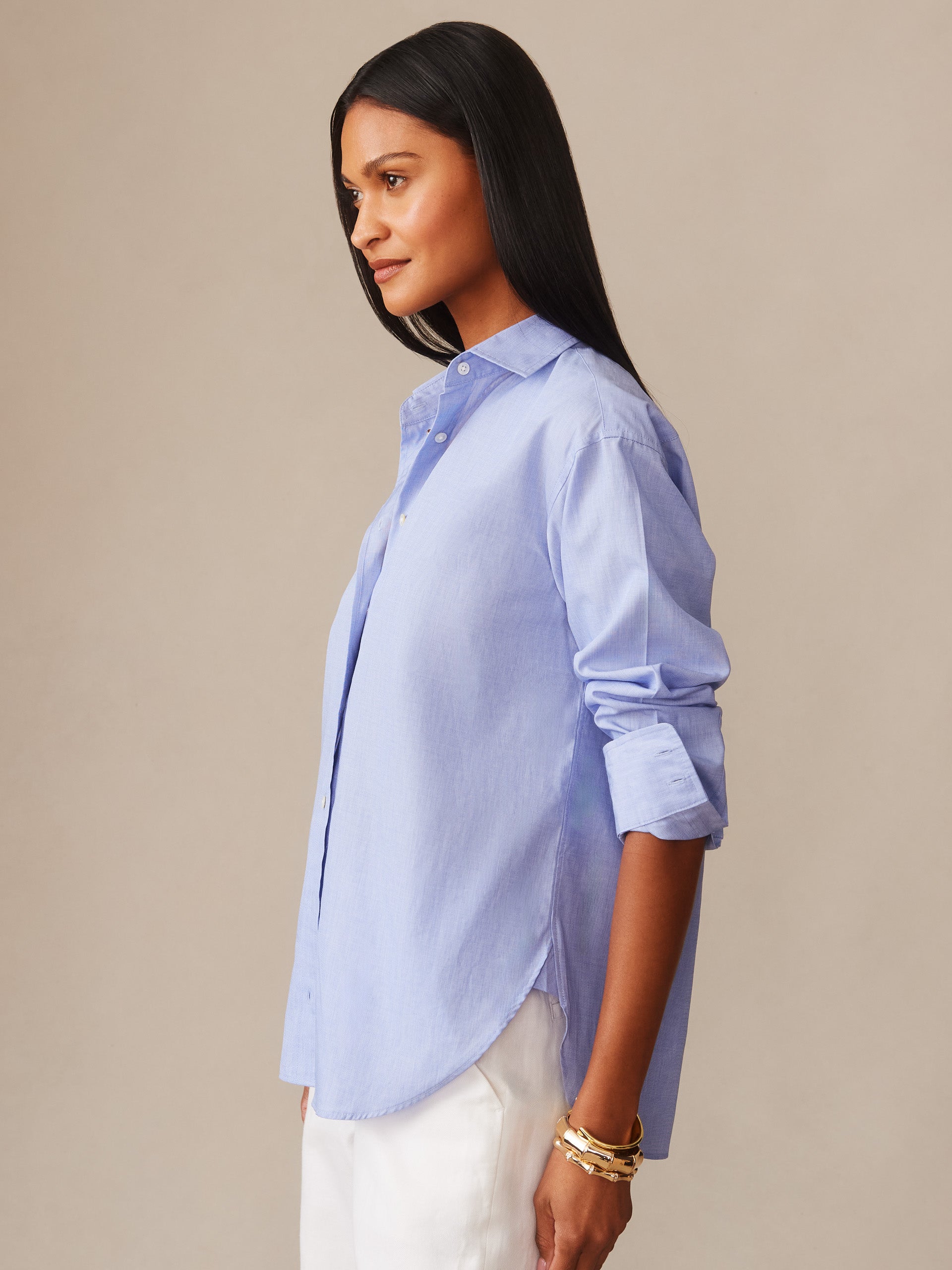 A woman wearing a light blue Finn Shirt by J.McLaughlin with rolled-up sleeves, paired with white pants and gold bracelets, showcasing effortlessly stylish, enduring designs and innovative fabrics.