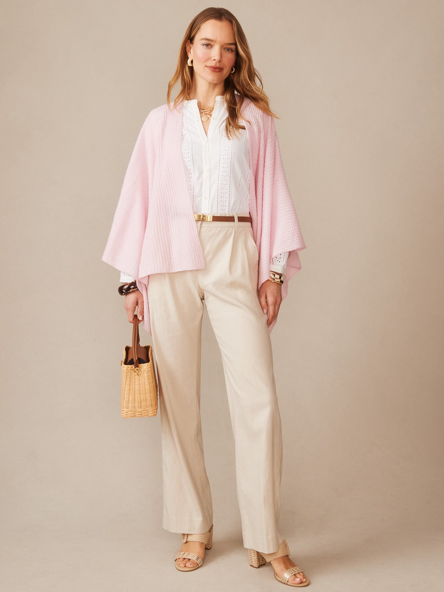Woman wearing a soft pink Julianne Cashmere Wrap over a white blouse and cream pants, accessorized with gold jewelry and a woven handbag, showcasing effortlessly stylish, enduring designs by J.McLaughlin.