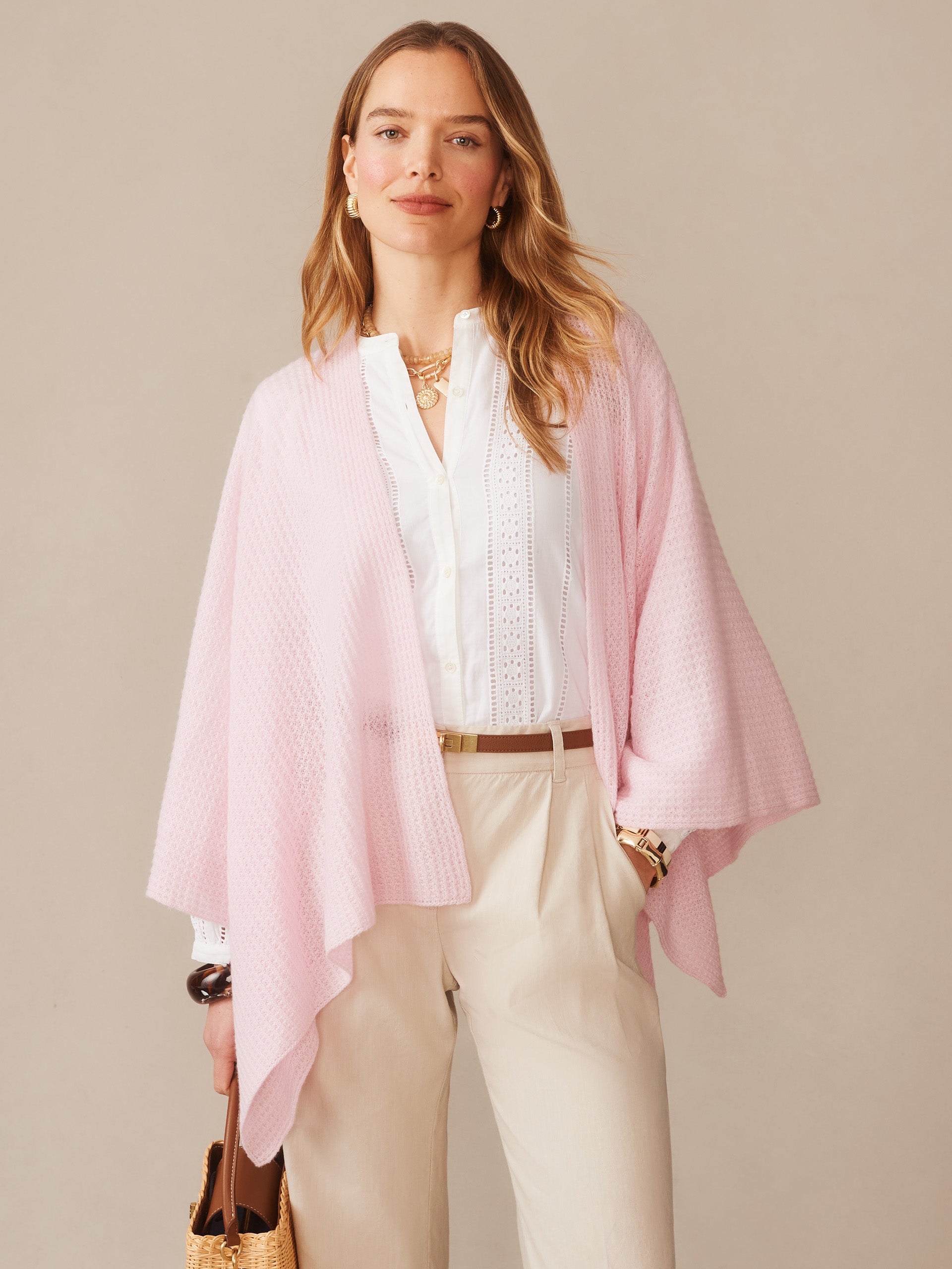Woman wearing a soft pink Julianne Cashmere Wrap over a white blouse and beige pants, styled with gold accessories and a woven handbag, embodying effortlessly stylish, enduring designs by J.McLaughlin.