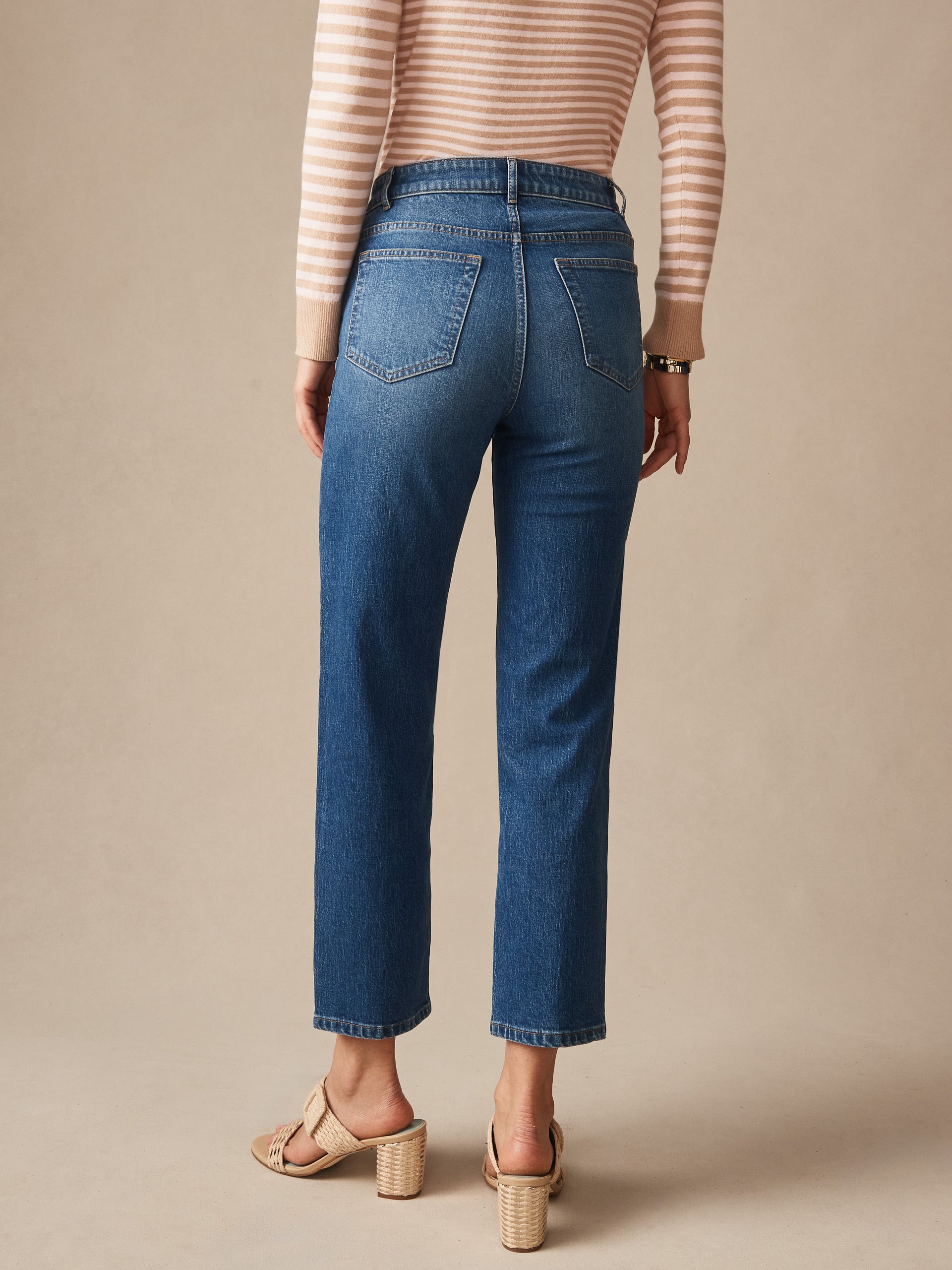 A rear view of a woman wearing Galveston Jeans by J.McLaughlin, paired with a striped top and woven heels, showcasing effortlessly stylish denim with enduring designs and a legacy of timeless fashion. A destination for defining style. Life, Well-Lived.