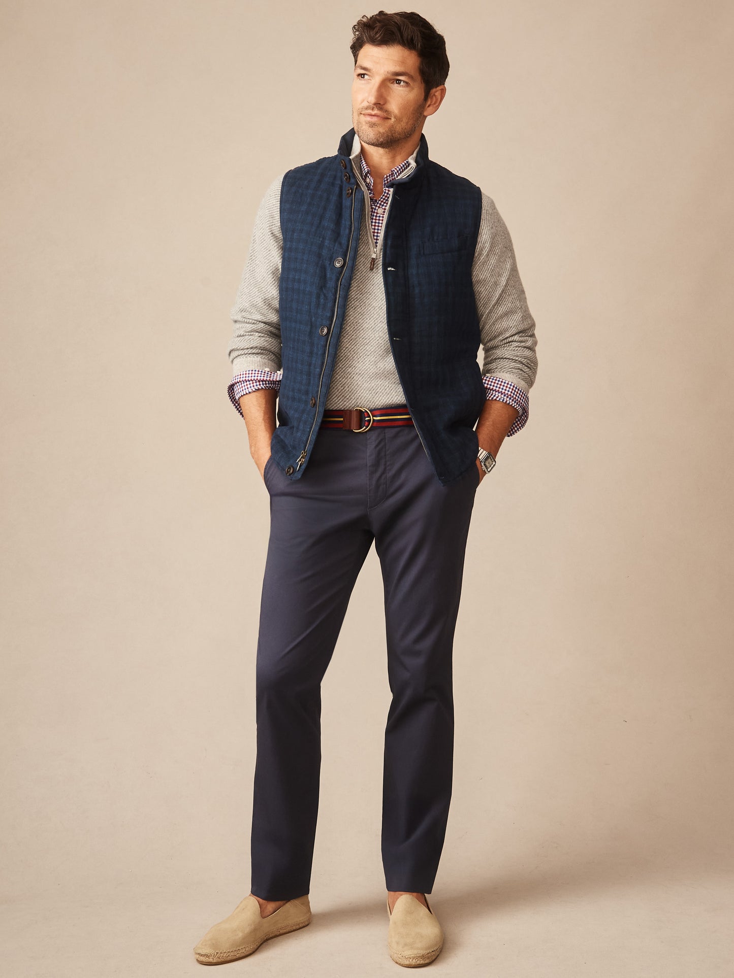Man wearing a navy shadow plaid quilted vest layered over a light gray sweater and checkered shirt, paired with navy pants and beige slip-on shoes, styled by J.McLaughlin.