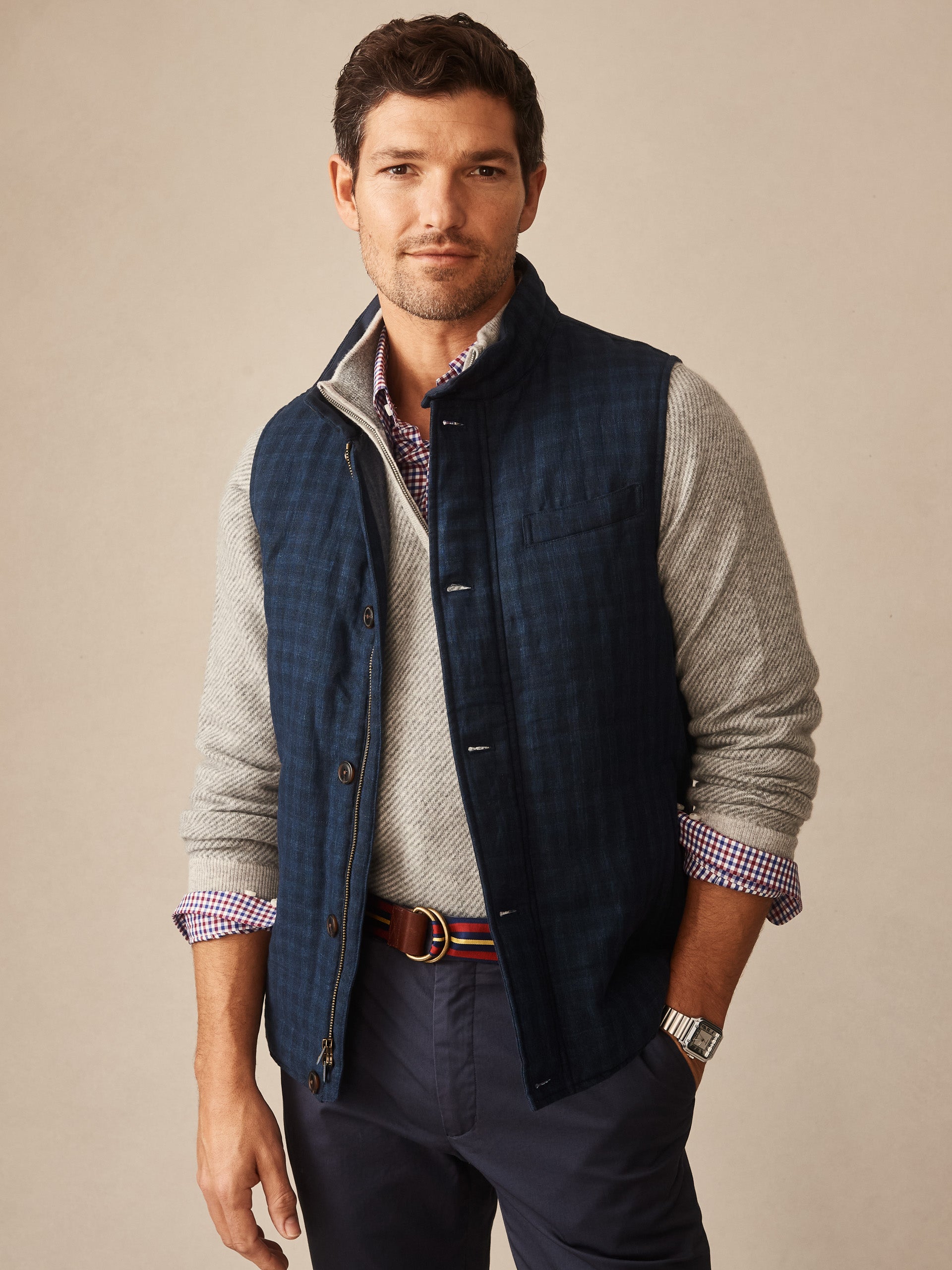 Man wearing a navy Game Quilted Vest in Shadow Plaid layered over a gray sweater and checkered shirt, styled with navy pants and a striped belt, by J.McLaughlin.