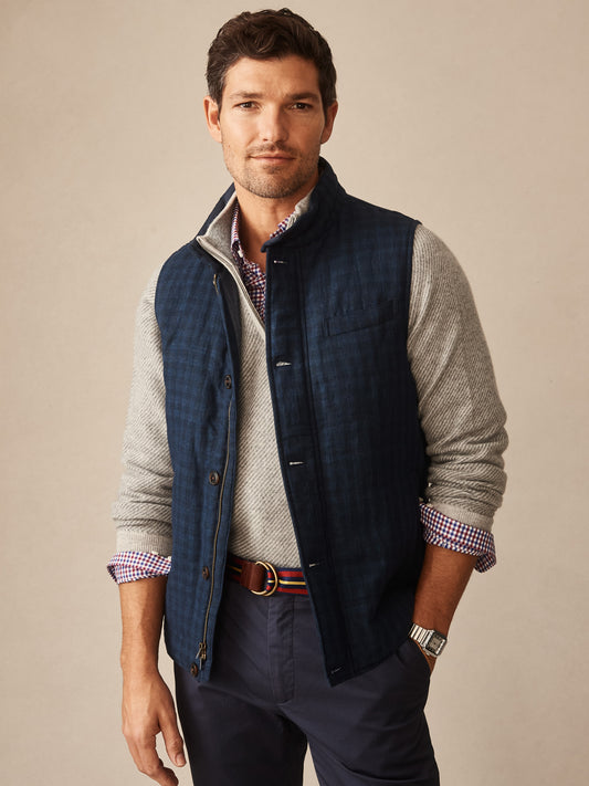 Man wearing a navy Game Quilted Vest in Shadow Plaid layered over a gray sweater and checkered shirt, styled with navy pants and a striped belt, by J.McLaughlin.