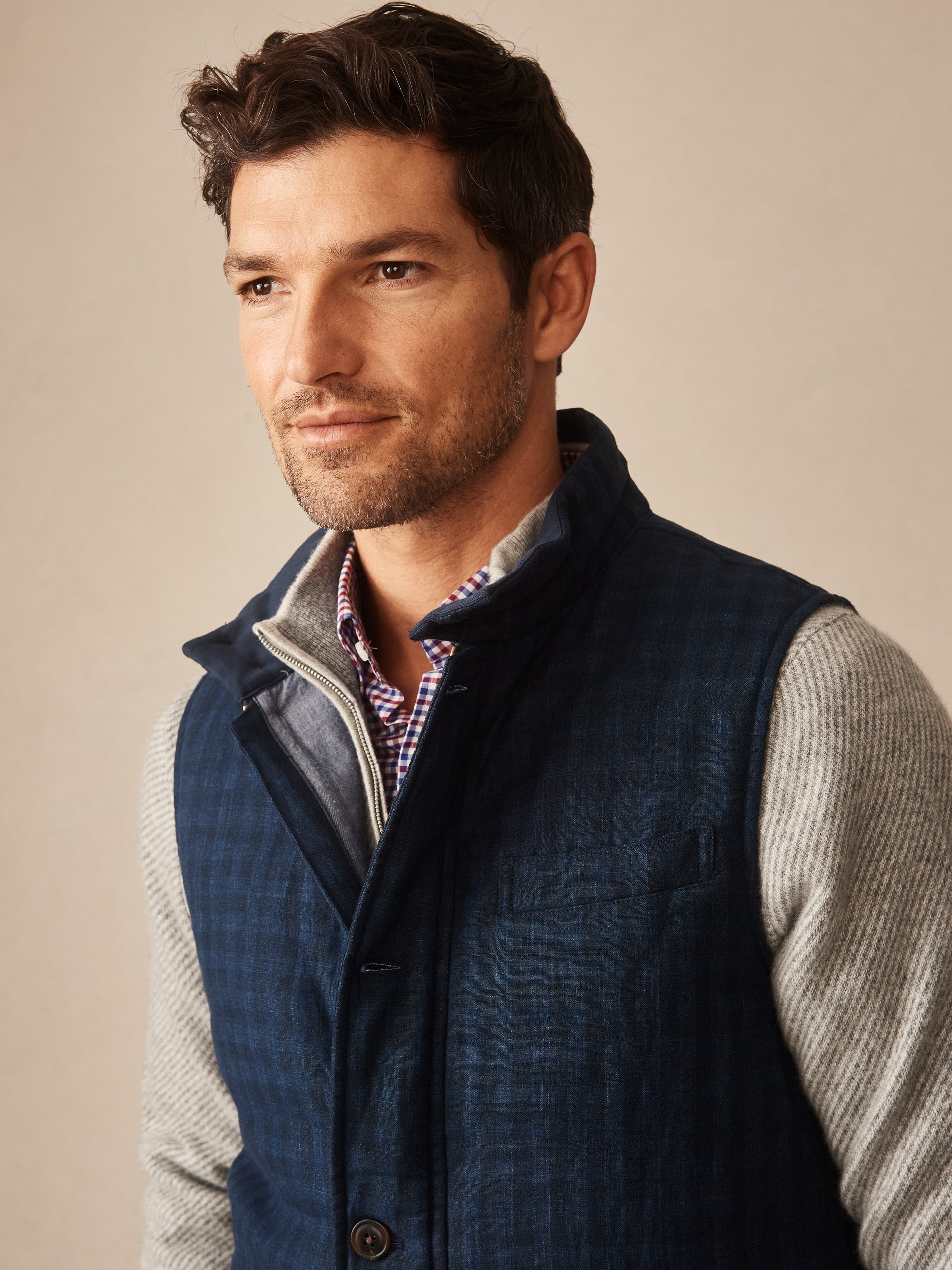 Man wearing a J.McLaughlin Game Quilted Vest in Shadow Plaid layered over a gray sweater and a checkered shirt, showcasing timeless, classic style.