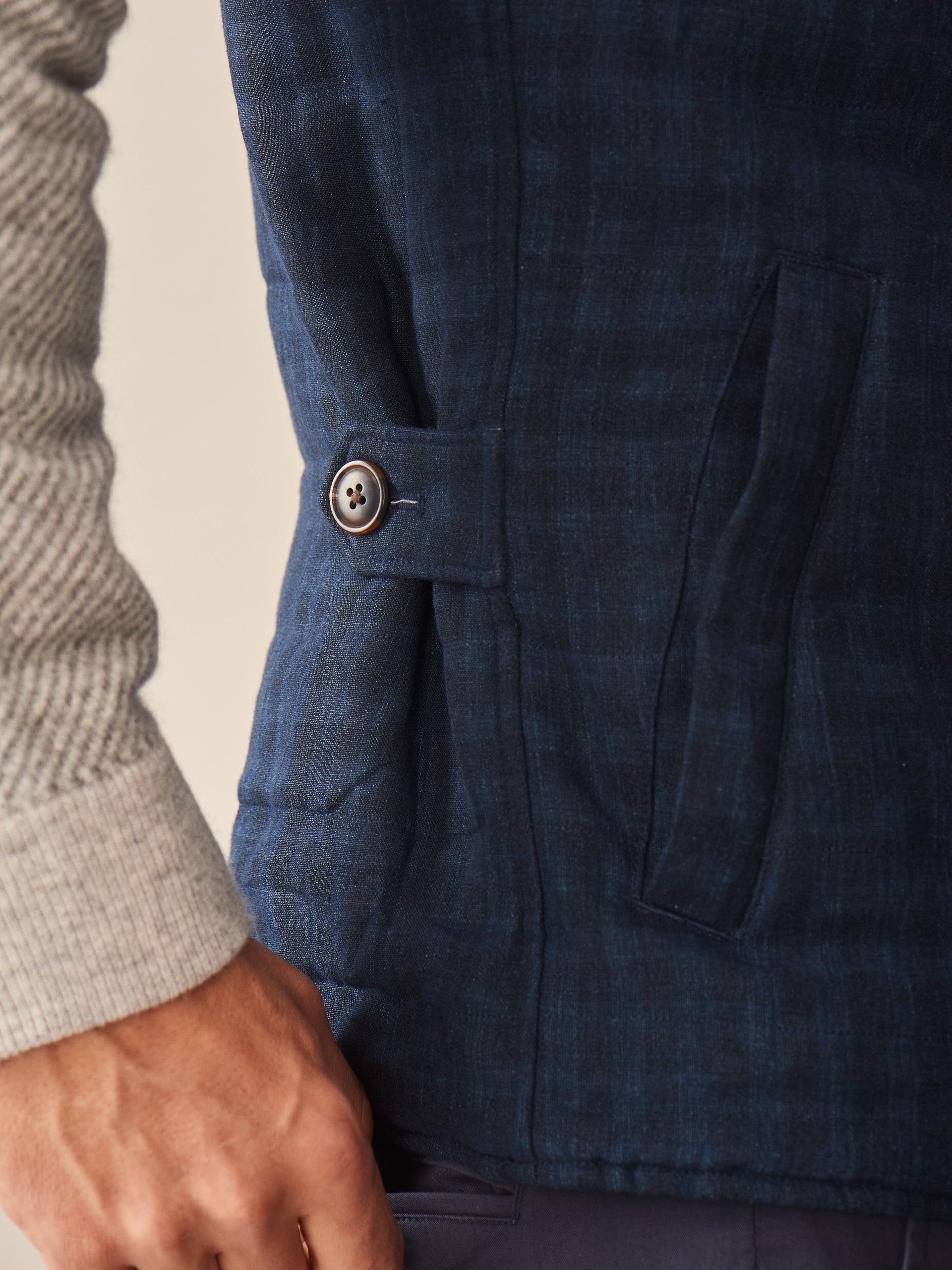 Close-up of a person wearing the Game Quilted Vest in Shadow Plaid by J.McLaughlin, showcasing the navy plaid pattern, side button detail, and textured pocket against a beige sweater sleeve.