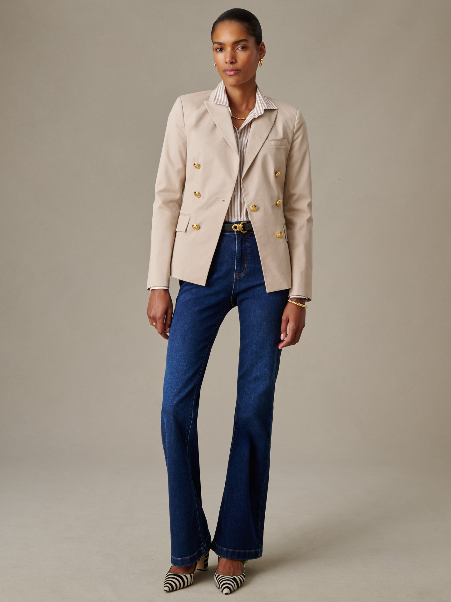 Woman wearing a beige Genevieve Cotton Twill Blazer by J.McLaughlin over a striped shirt, paired with dark blue jeans and striped pointed-toe shoes, showcasing effortless style.