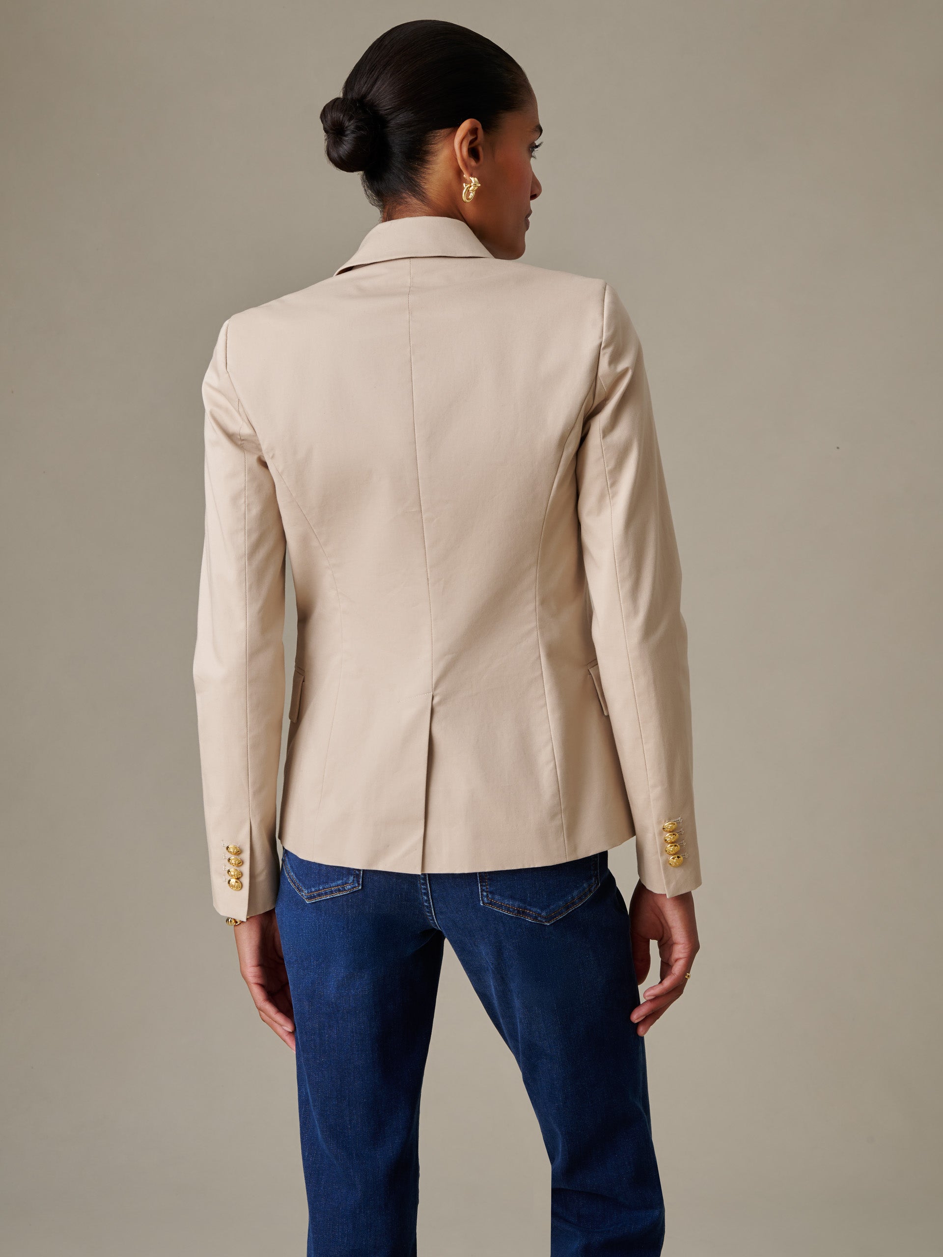 A woman wearing a beige Genevieve Cotton Twill Blazer by J.McLaughlin, styled with dark blue jeans, showcasing the blazer’s tailored back and gold button details on sleeves.