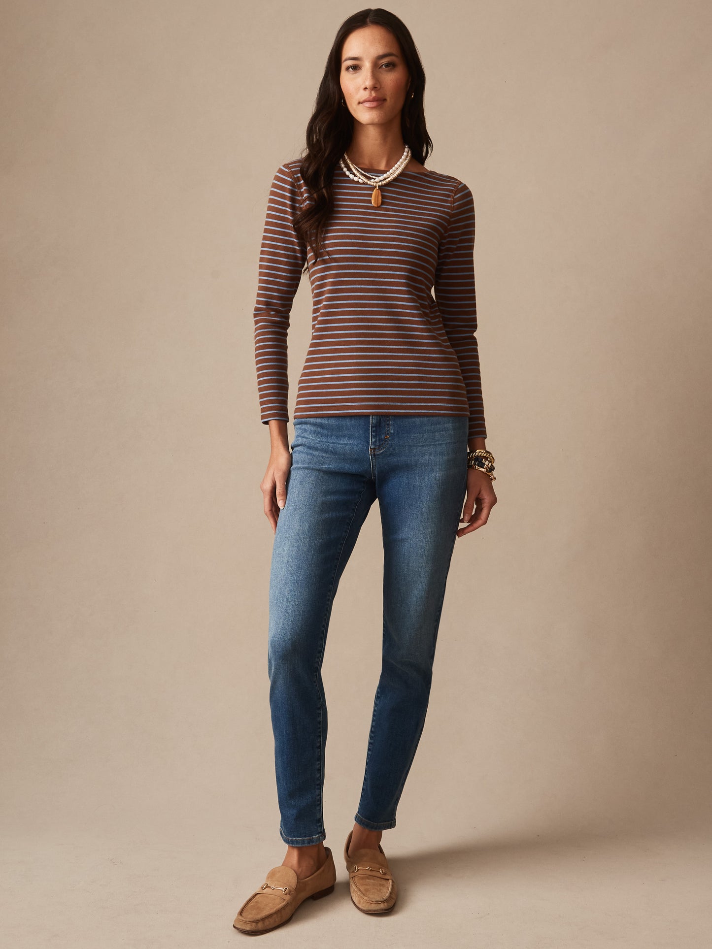 Woman wearing a brown and blue striped long-sleeve top paired with blue jeans and tan loafers, showcasing the Goldie Tee in Stripe by J.McLaughlin, combining effortlessly stylish and enduring designs with innovative fabrics for a legacy of timeless fashion. A destination for defining style. Life, Well-Lived.