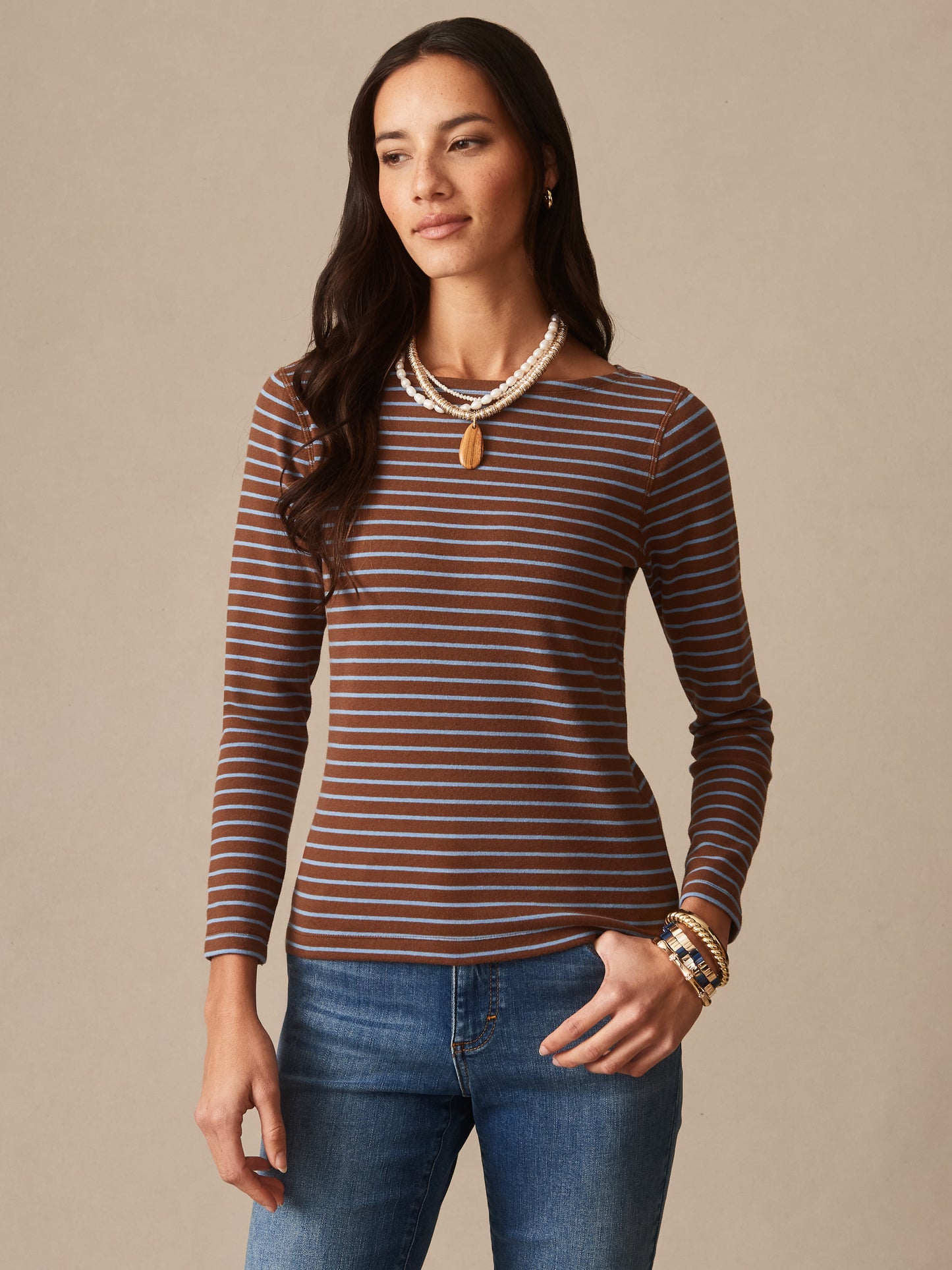 Woman wearing a brown and blue striped Goldie Tee in Stripe by J.McLaughlin paired with jeans and layered necklaces, showcasing effortlessly stylish, enduring designs in innovative fabrics.