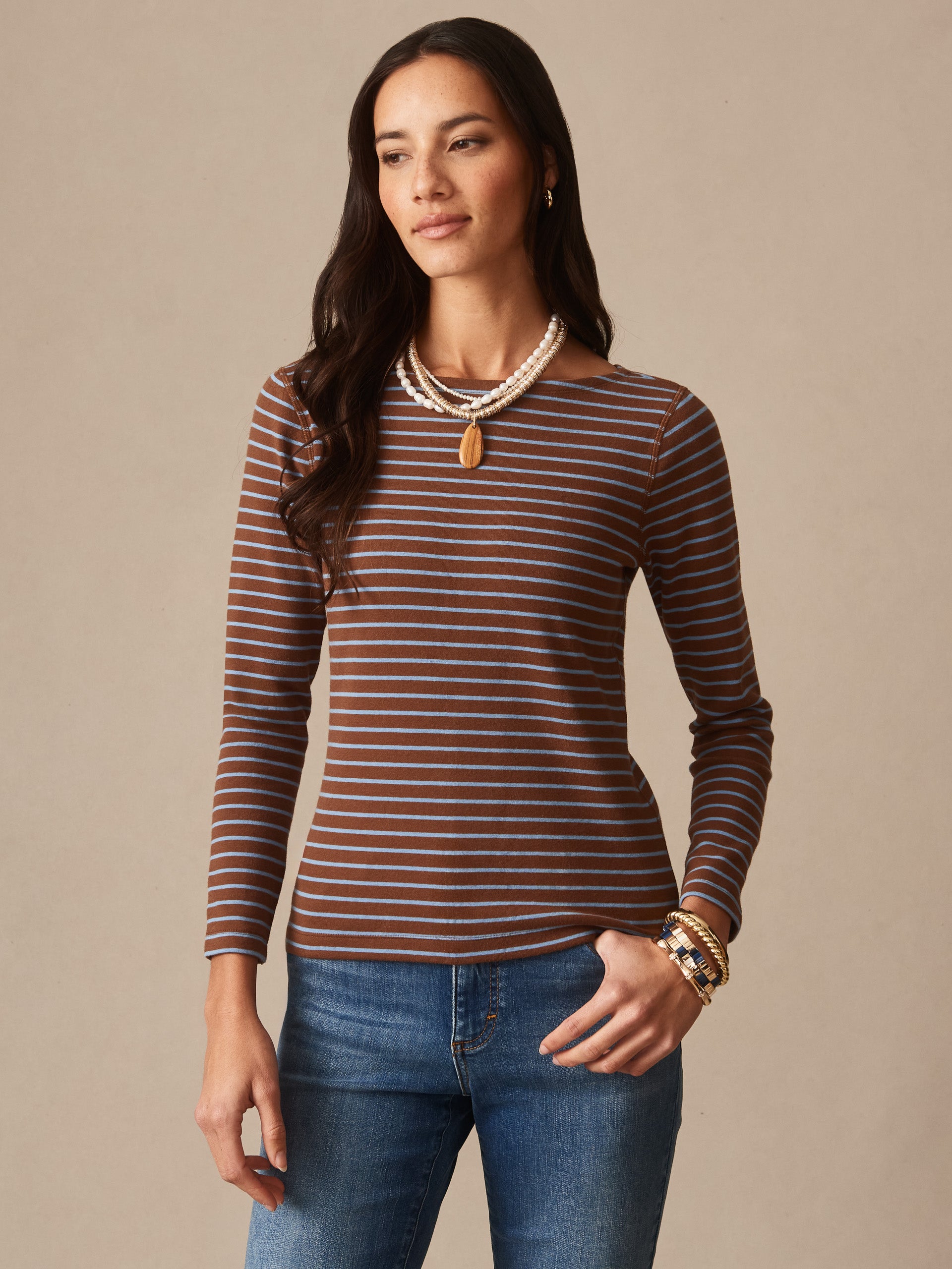 Woman wearing a brown and blue striped Goldie Tee in Stripe by J.McLaughlin paired with jeans and layered necklaces, showcasing effortlessly stylish, enduring designs in innovative fabrics.
