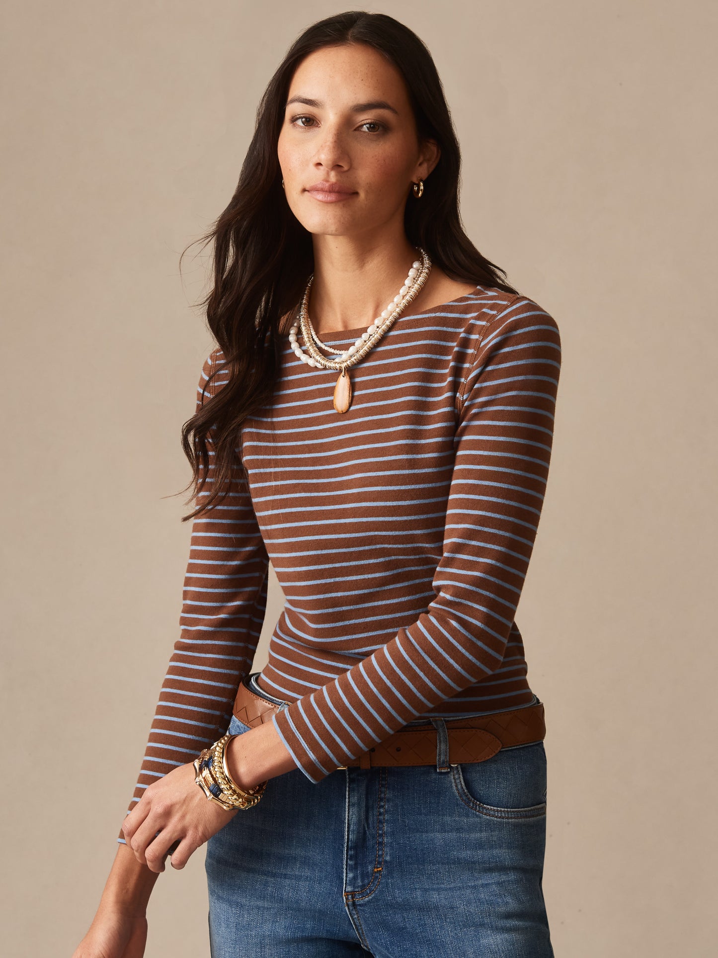 Woman wearing a brown and light blue striped Goldie Tee in Stripe by J.McLaughlin paired with denim jeans and layered beaded necklaces, showcasing effortlessly stylish, enduring designs with innovative fabrics. A destination for defining style. Life, Well-Lived.