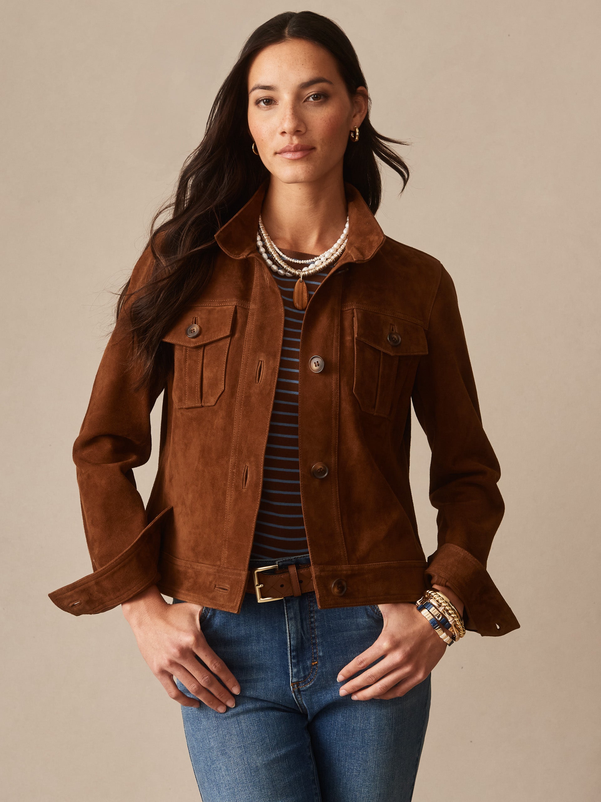 Woman wearing a brown Frances Suede Jacket by J.McLaughlin over a striped top and jeans, accessorized with layered necklaces and bracelets, showcasing effortlessly stylish, enduring designs.