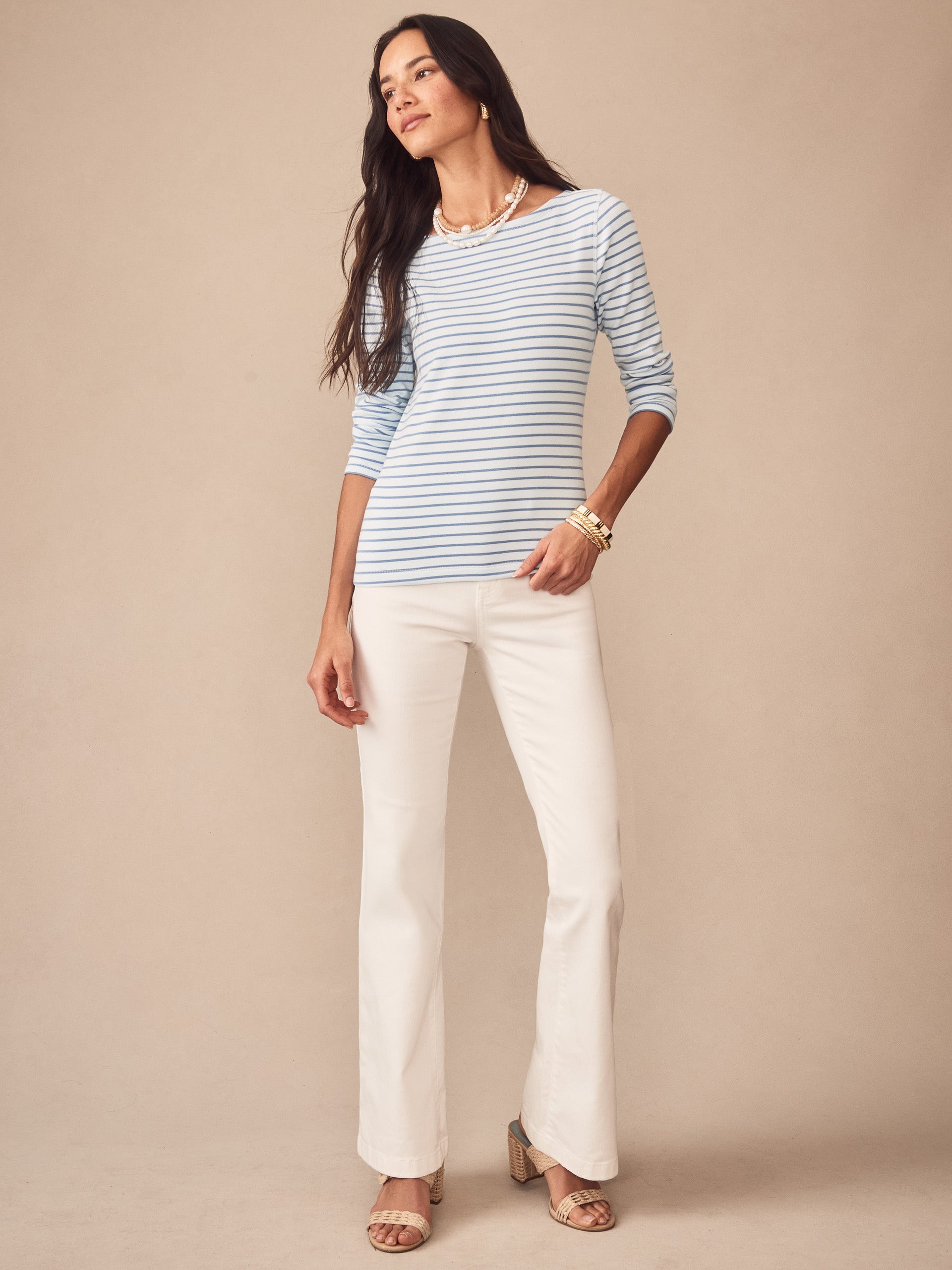 A woman models the Goldie Tee in Stripe by J.McLaughlin, featuring light blue and white stripes with three-quarter sleeves, paired with white pants and gold accessories, embodying effortlessly stylish, enduring designs and innovative fabrics, a legacy of timeless fashion at a destination for defining style. Life, Well-Lived.
