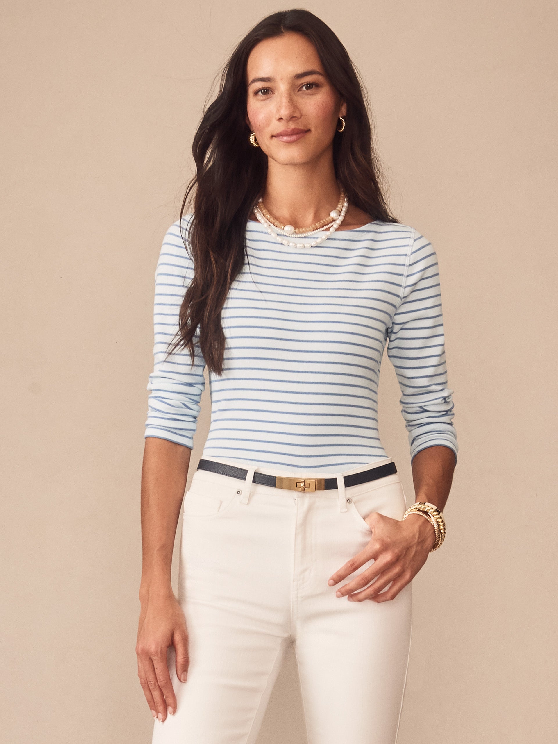 Woman wearing a Goldie Tee in Stripe with light blue and white stripes, paired with white pants and gold accessories, showcasing J.McLaughlin’s effortlessly stylish and enduring designs, a destination for defining style. Life, Well-Lived.