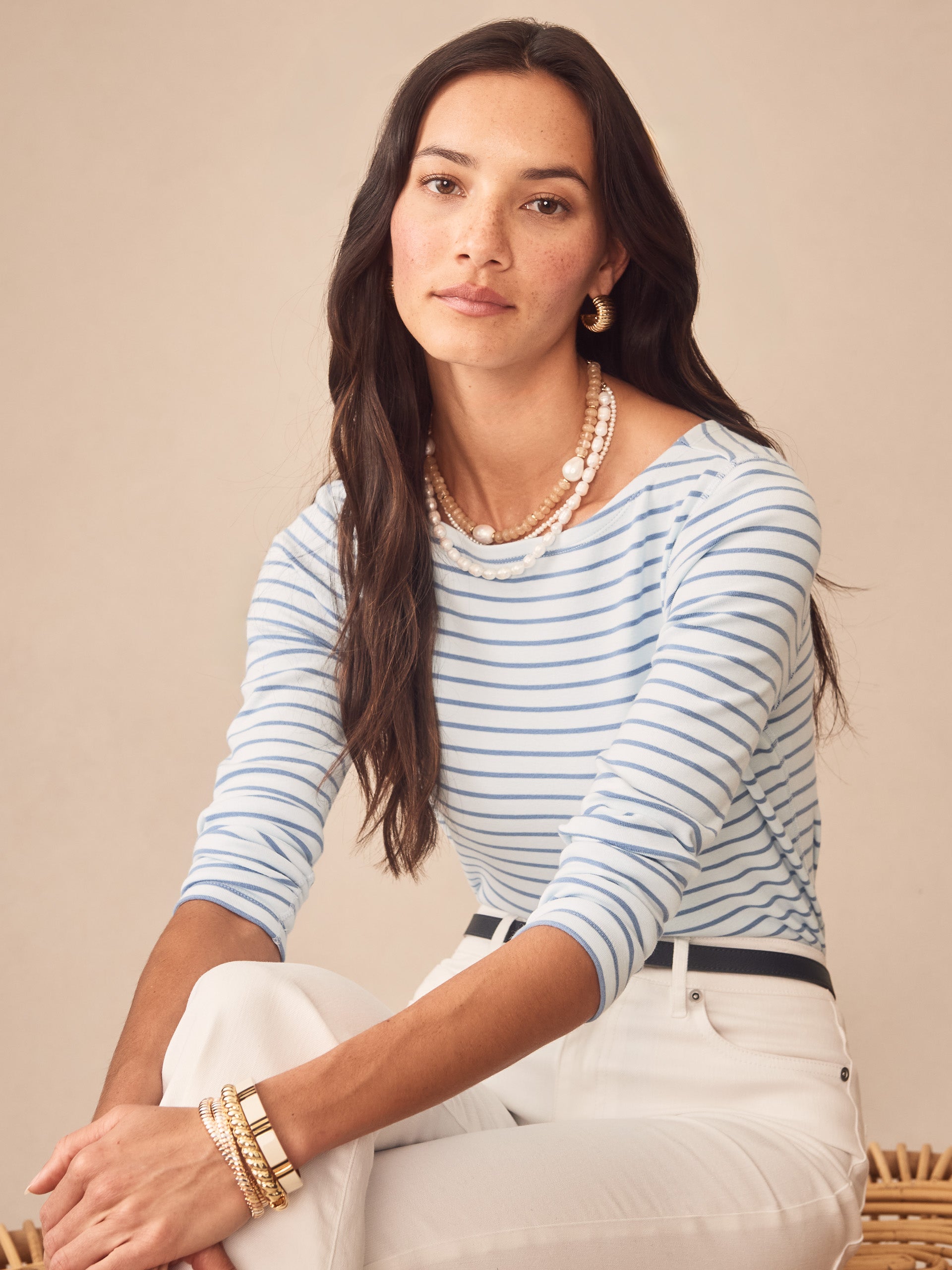 Woman wearing the Goldie Tee in Stripe by J.McLaughlin, featuring soft blue stripes and a classic fit, paired with white pants and layered pearl necklaces, embodying effortlessly stylish and enduring designs in innovative fabrics for a legacy of timeless fashion at a destination for defining style. Life, Well-Lived.