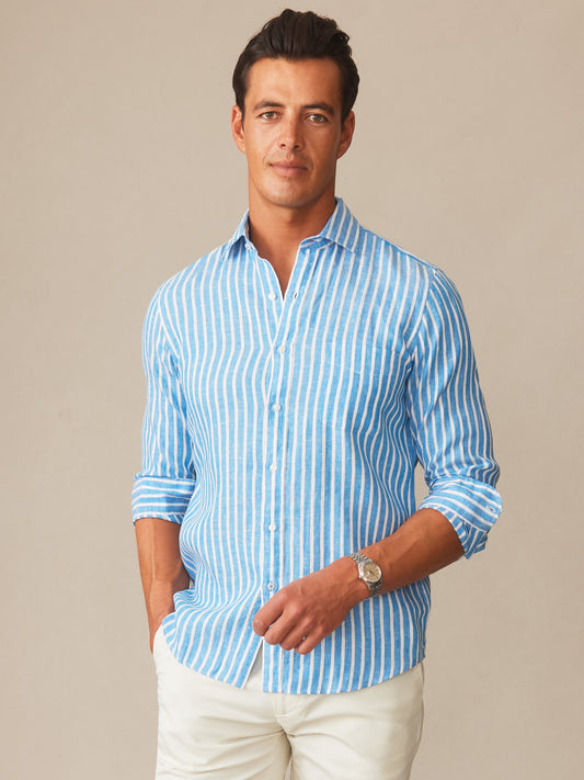 Man wearing a blue and white striped Gramercy Linen Shirt in Dobby Stripe by J.McLaughlin, styled casually with rolled sleeves and white pants, showcasing effortlessly stylish, enduring designs with innovative fabrics, a destination for defining style. Life, Well-Lived.
