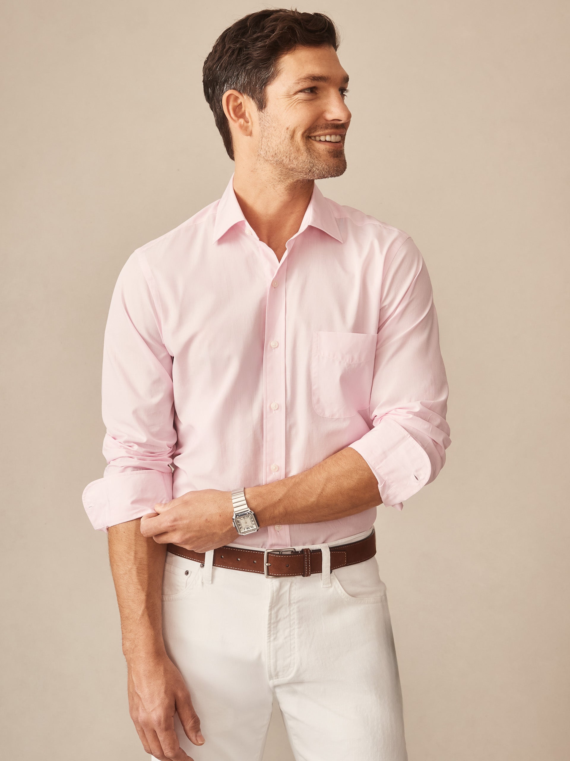 A smiling man wearing a light pink Gramercy Cotton Shirt by J.McLaughlin with rolled-up sleeves, paired with white pants and a brown belt, showcasing classic style and craftsmanship.