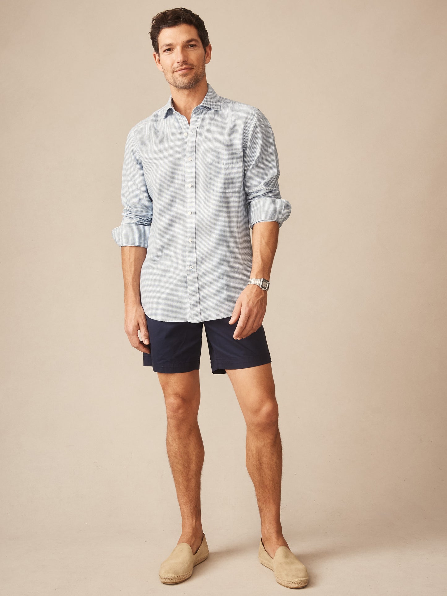 Man wearing a Gramercy Linen Shirt in Micro Gingham by J.McLaughlin in light blue with rolled sleeves, paired with navy shorts and beige slip-on shoes, embodying effortlessly stylish, enduring designs.