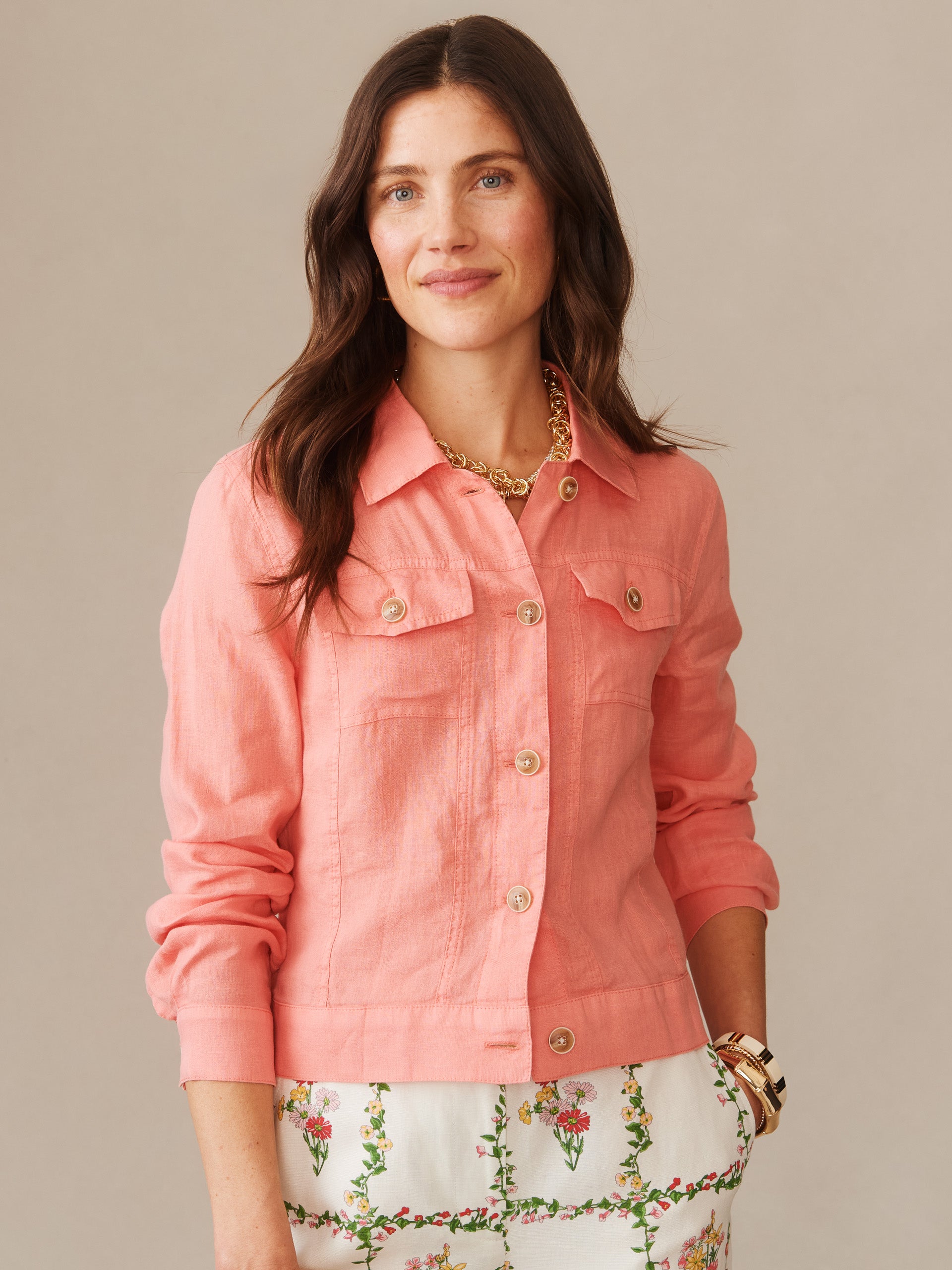 A woman wearing the effortlessly stylish Halo Linen Jacket in coral by J.McLaughlin, paired with floral patterned pants and gold accessories, showcasing enduring designs and legacy of timeless fashion.